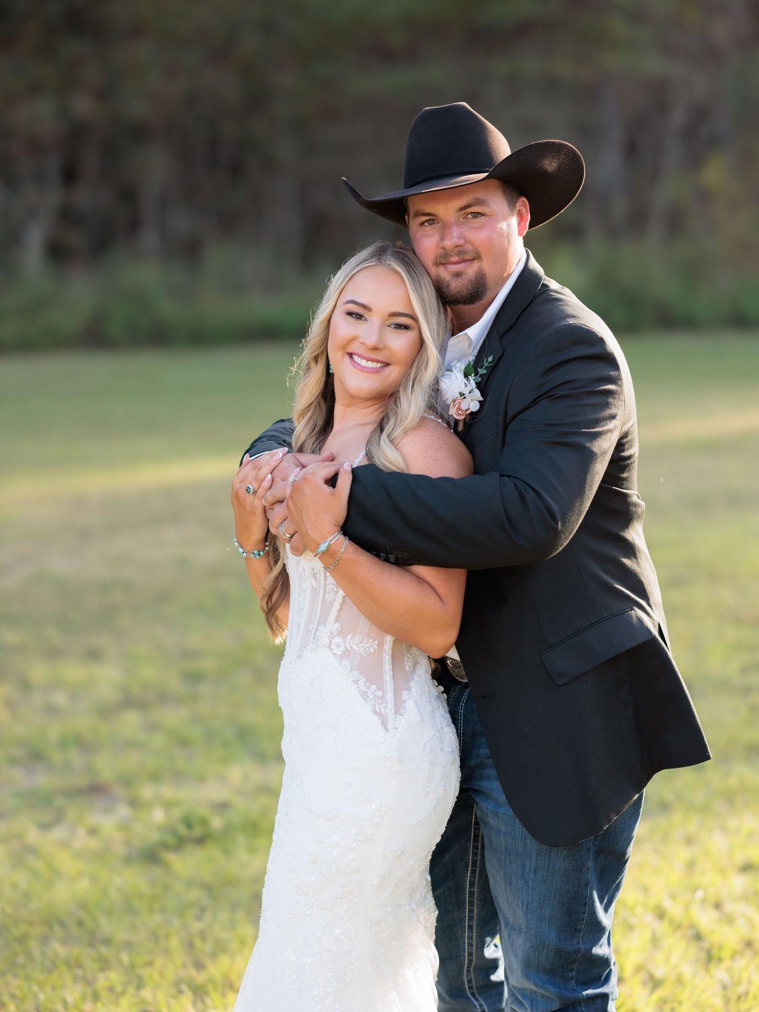 A couple in wedding attire embrace outdoors, with the groom wearing a black cowboy hat and suit jacket with jeans.
