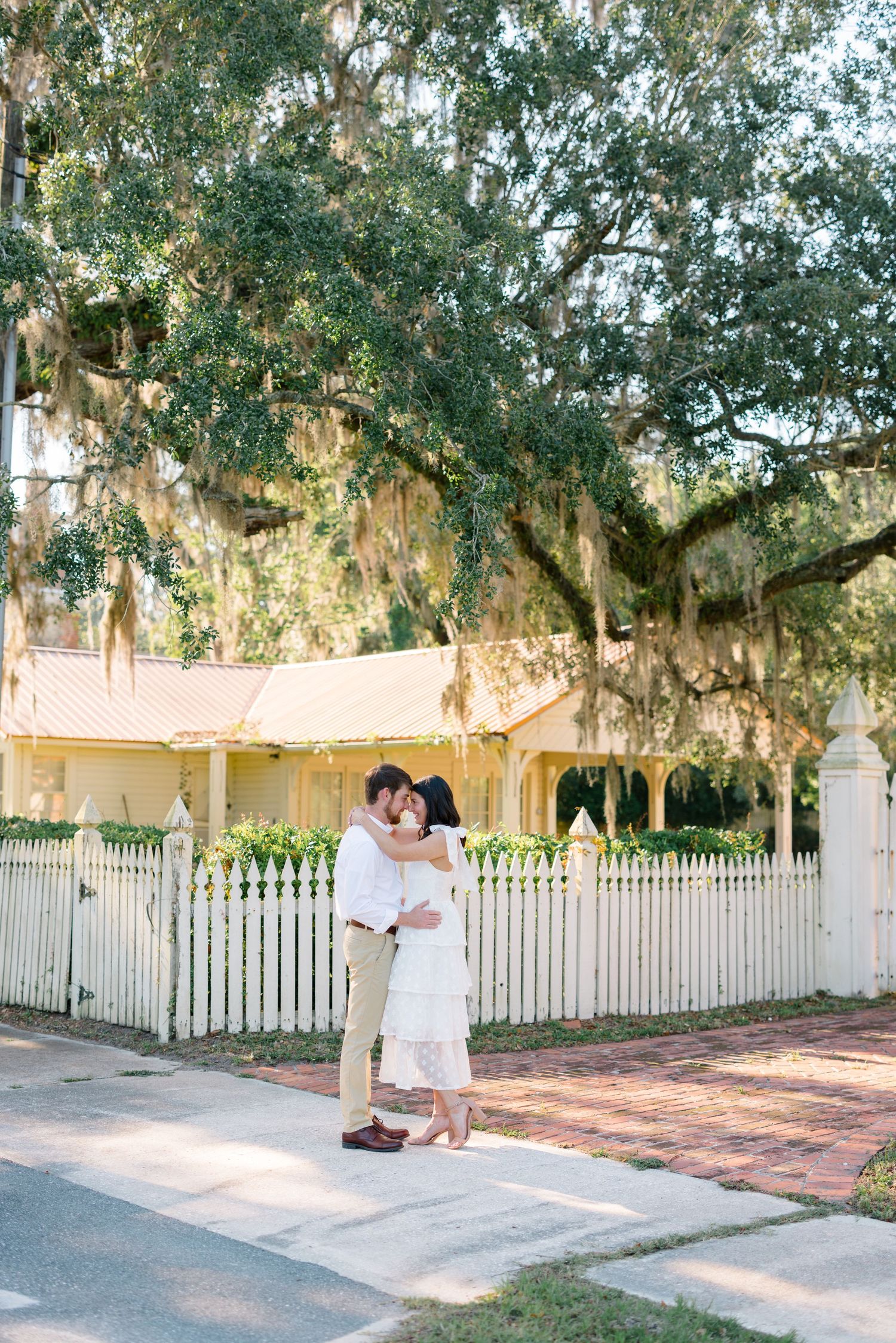 A couple embraces on a sidewalk in front of a yellow house with Spanish moss hanging from trees.