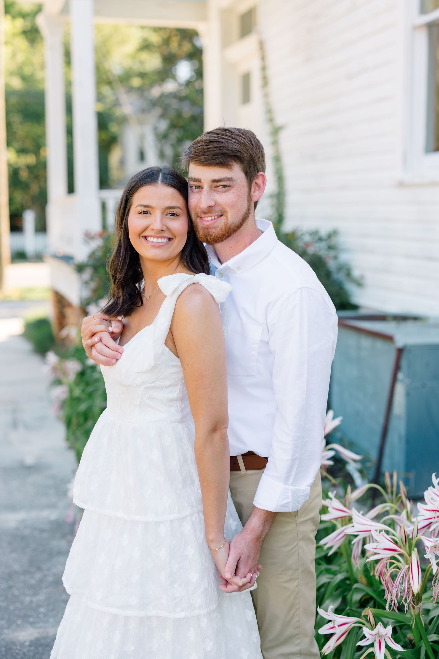 A couple poses together in coordinated white outfits near a garden with pink flowers.
