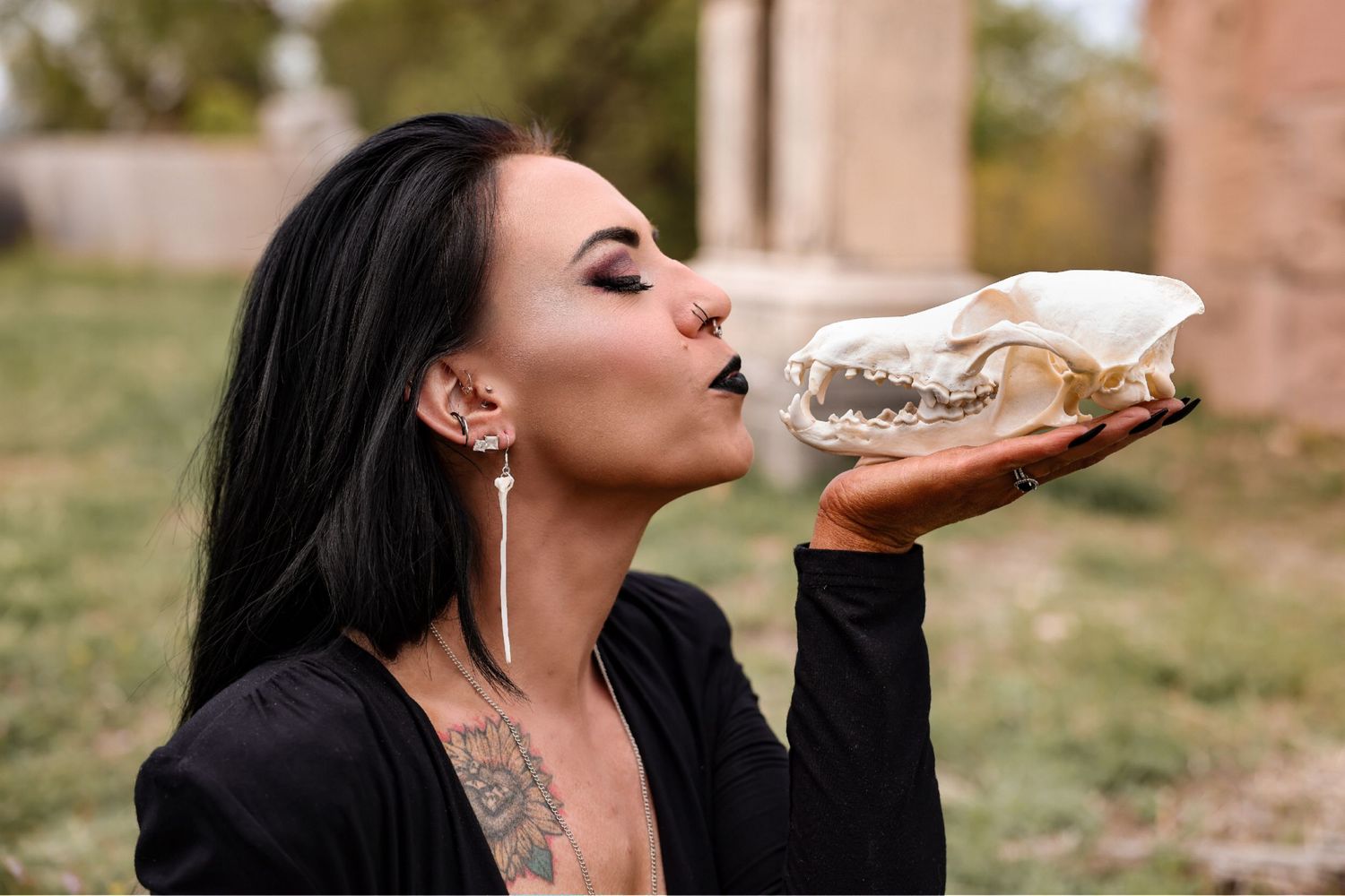 Person with black hair and tattoos kissing an animal skull outdoors.