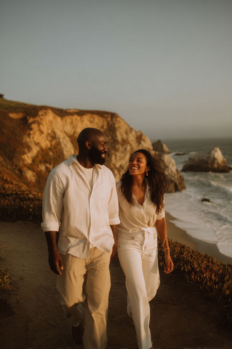 Couple in white outfits walking along a coastal cliff at golden hour, smiling and enjoying a romantic beach stroll.