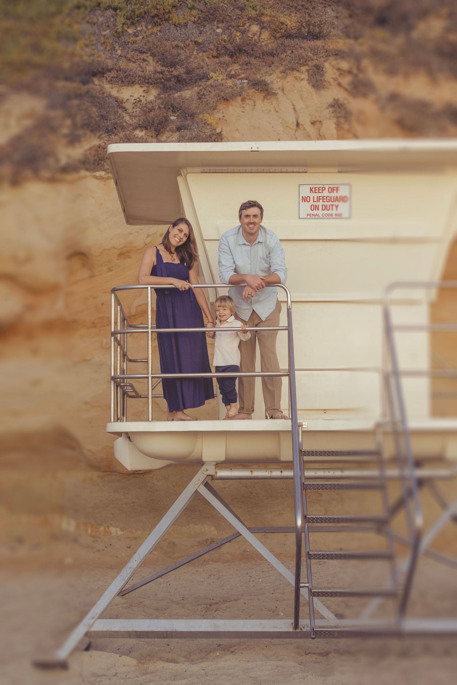 Family of three posing on a lifeguard tower at the beach, with sandy cliffs in the background.