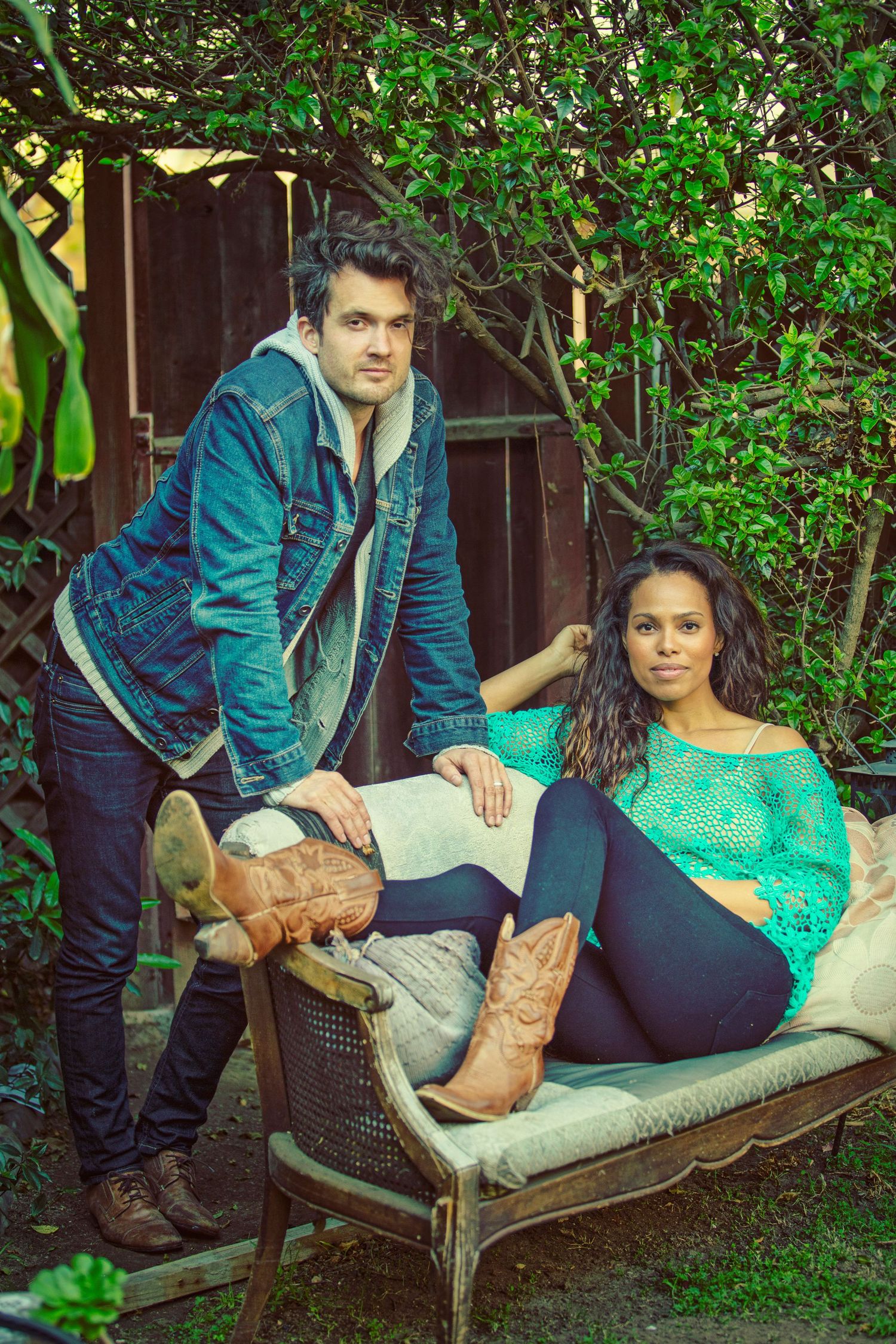 A man in a denim jacket stands beside a woman in a teal top sitting on a vintage chair in a lush garden.