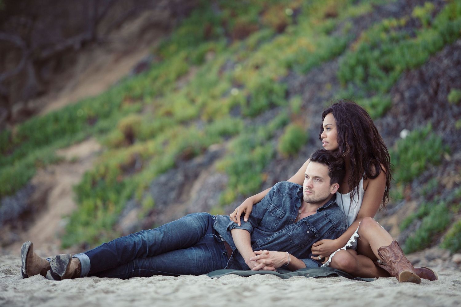Couple relaxing on sandy beach, man lying with head on woman's lap, enjoying a romantic outdoor moment.