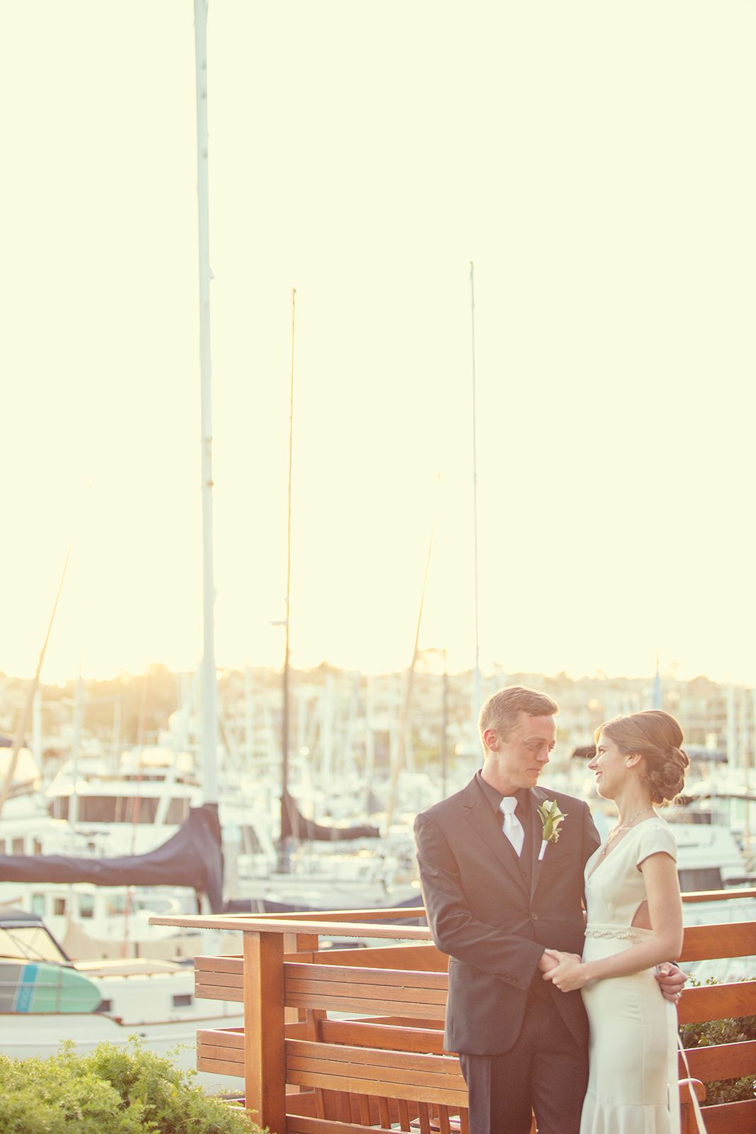 Bride and groom stand together on waterfront deck at sunset with sailboats and marina in the background.