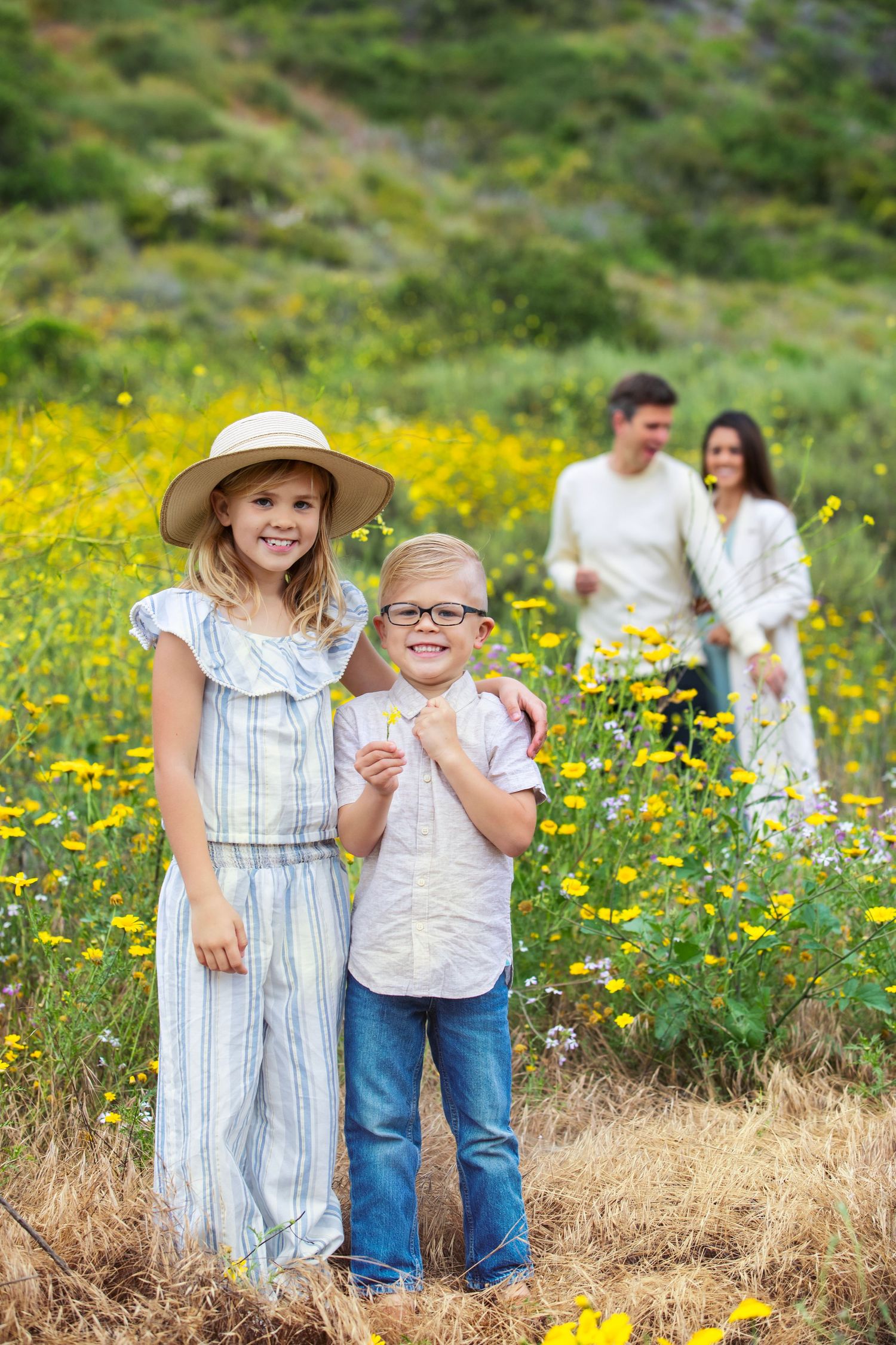 Two children smile in a sunny wildflower field while their parents stand together in the background.