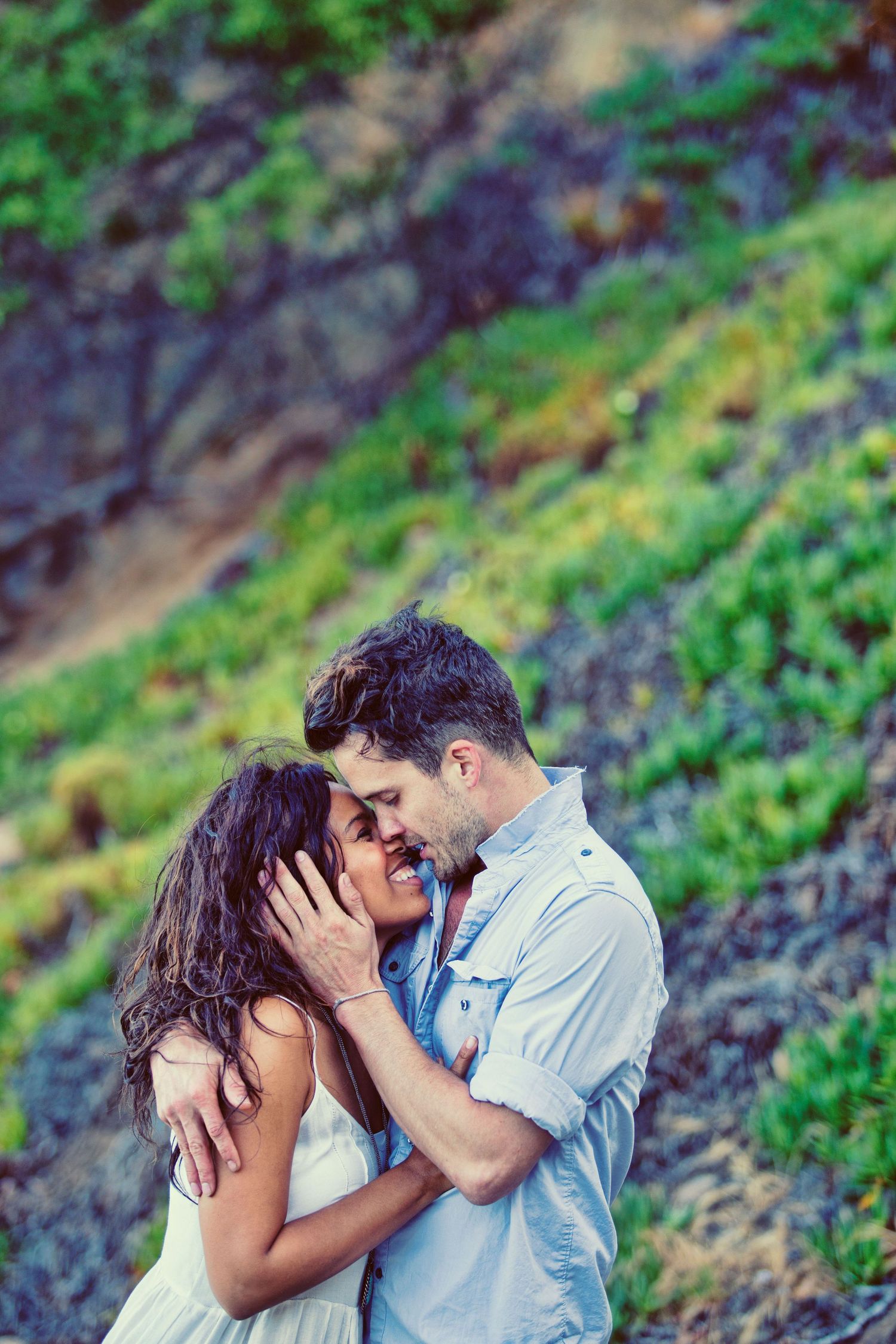A romantic couple embraces tenderly outdoors, with lush green hillside vegetation in the background.