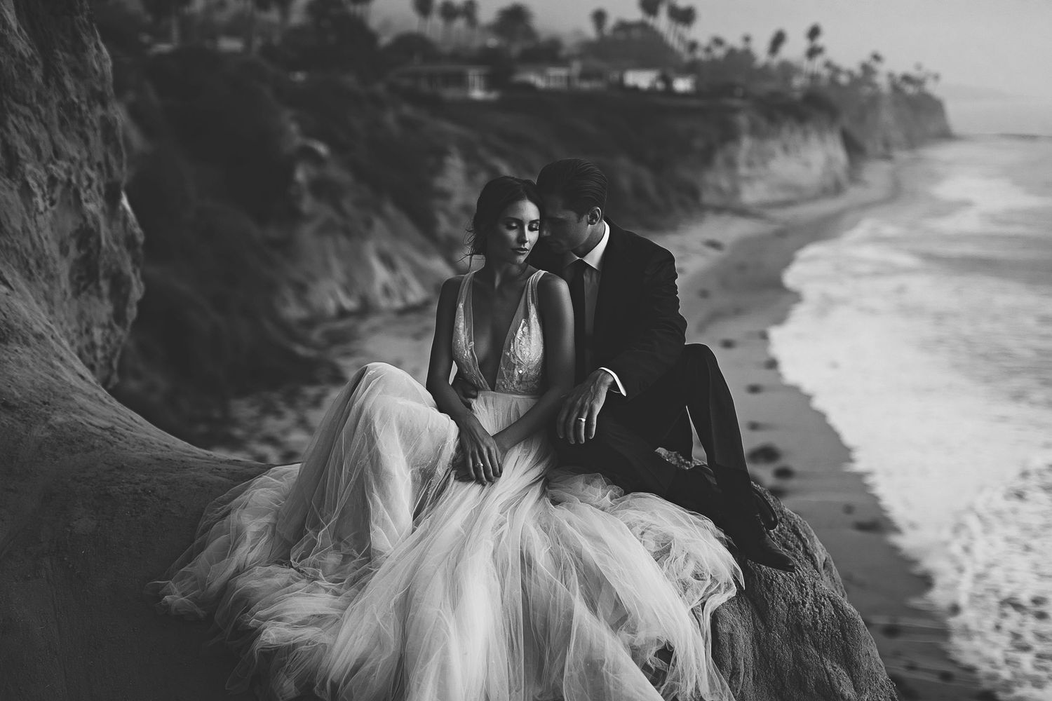 Black and white photo of a couple in wedding attire sitting on coastal cliffs with ocean waves in the background.