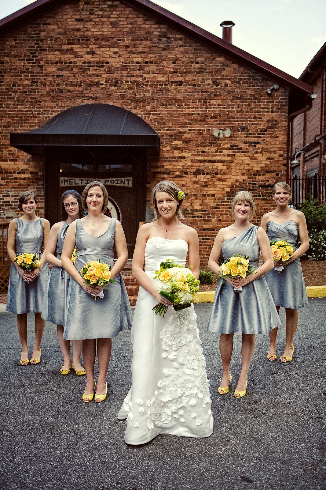 Bride in white gown with bridesmaids in silver dresses and yellow heels holding yellow floral bouquets outside brick building.