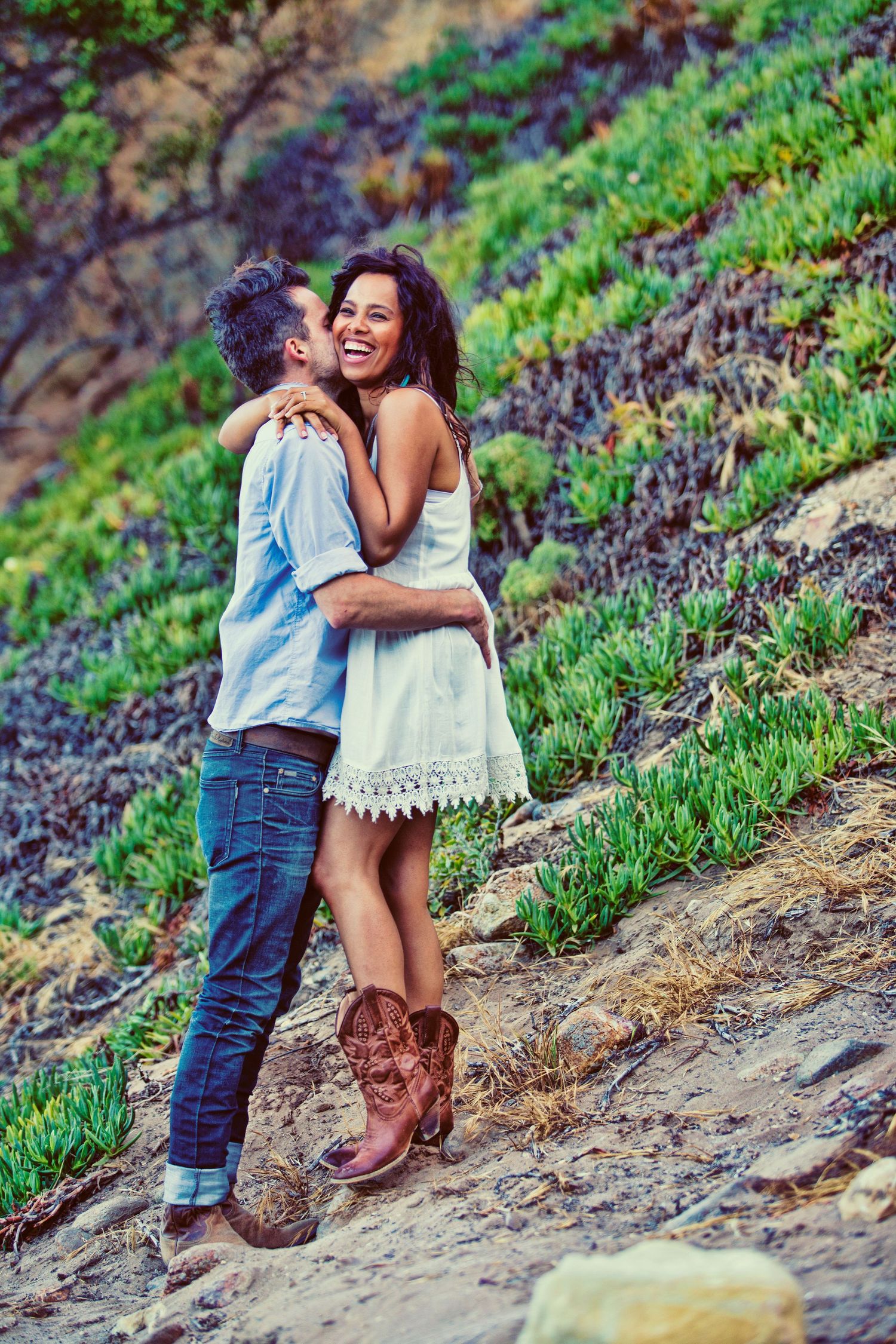 Happy couple embracing and laughing outdoors on a rocky hillside with green plants, woman in white dress and cowboy boots.