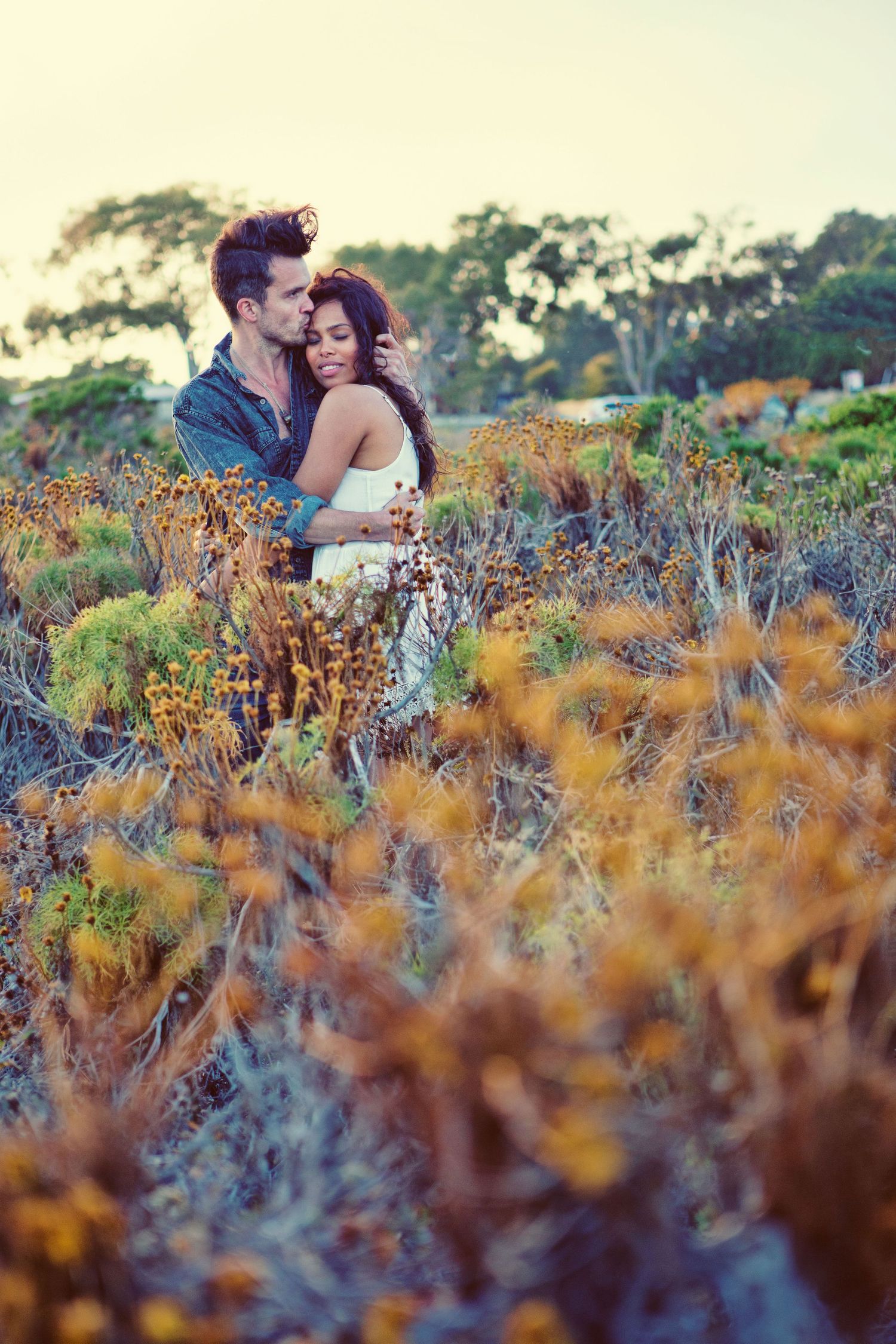 Romantic couple embracing in a field of golden wildflowers at sunset, surrounded by lush coastal vegetation.