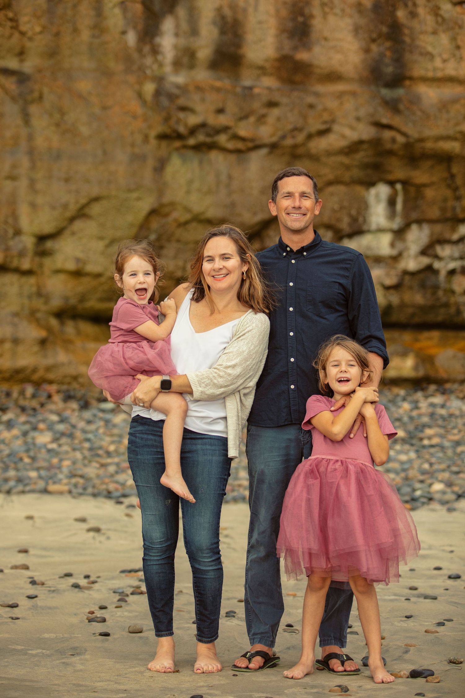 Family of four smiling on a rocky beach, two girls in pink dresses, parents in casual attire, with cliffs in the background.