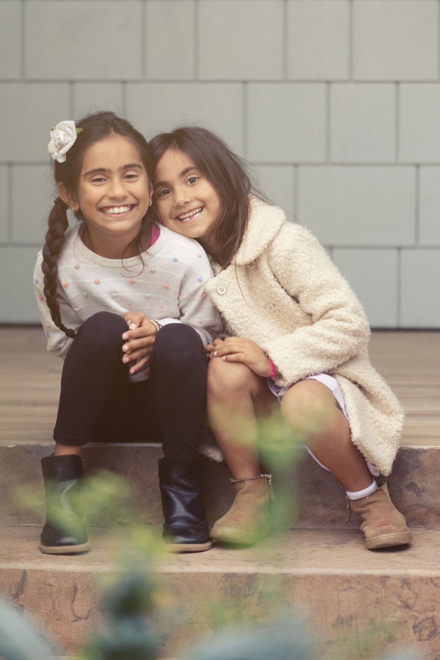 Two smiling girls in casual fall outfits sit together on steps, sharing a warm embrace.
