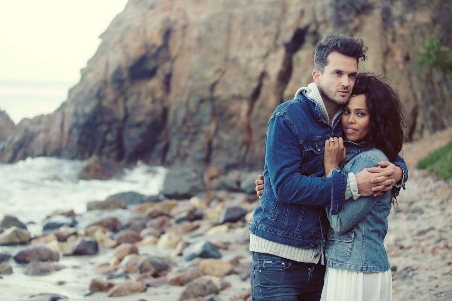 Couple embracing on a rocky beach shore, man in denim jacket holding woman in white skirt near ocean cliffs.