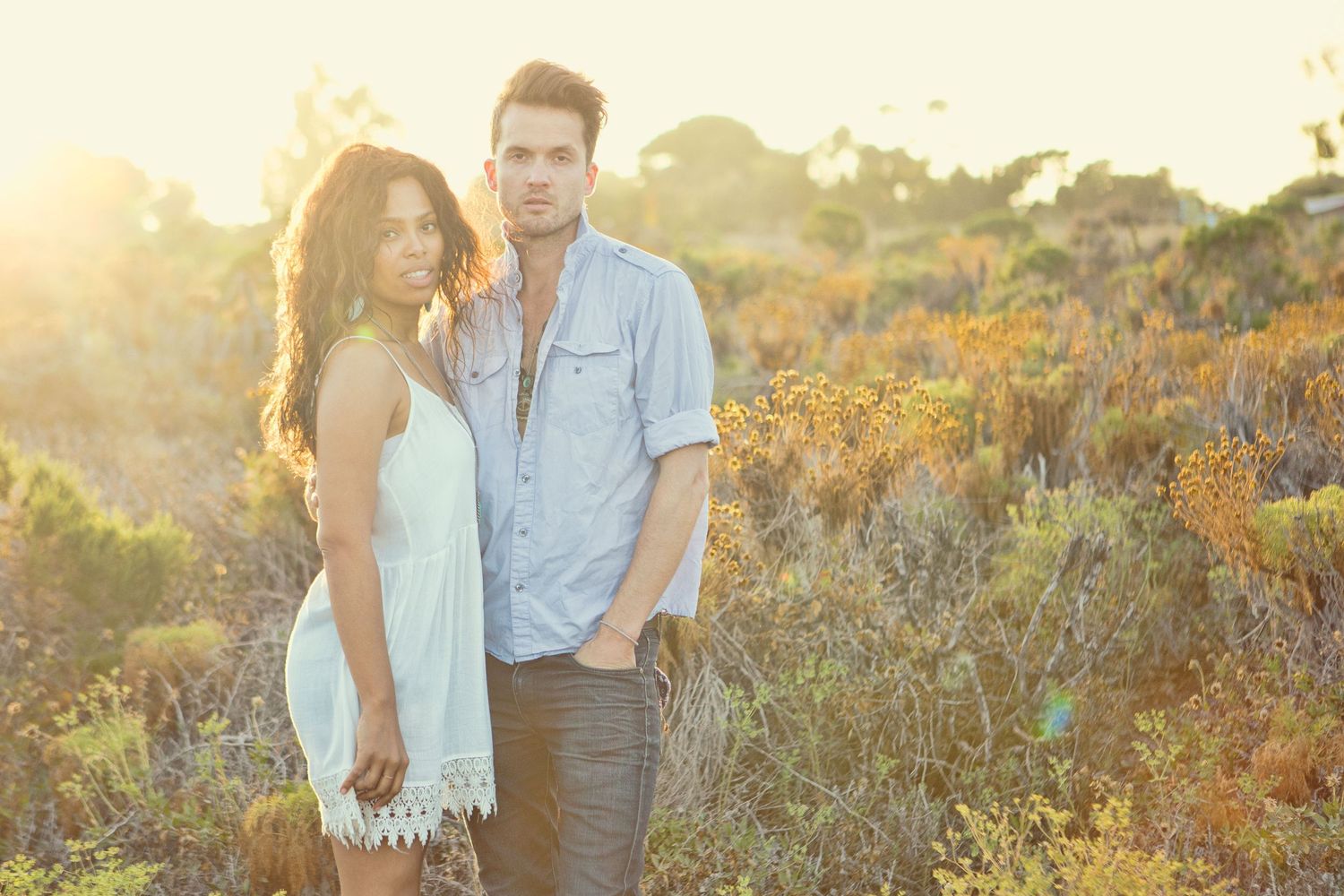 A young couple standing together in a sunlit field with golden hour light and wild shrubs in the background.