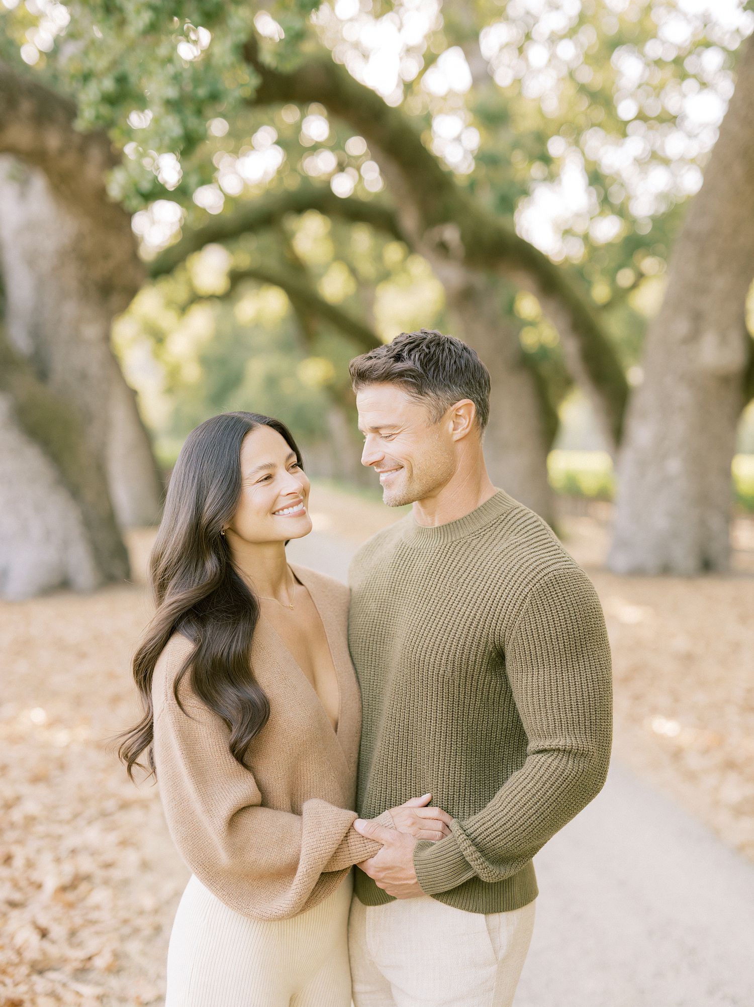 A couple smiles warmly at each other in cozy sweaters beneath oak trees during a romantic outdoor engagement session.