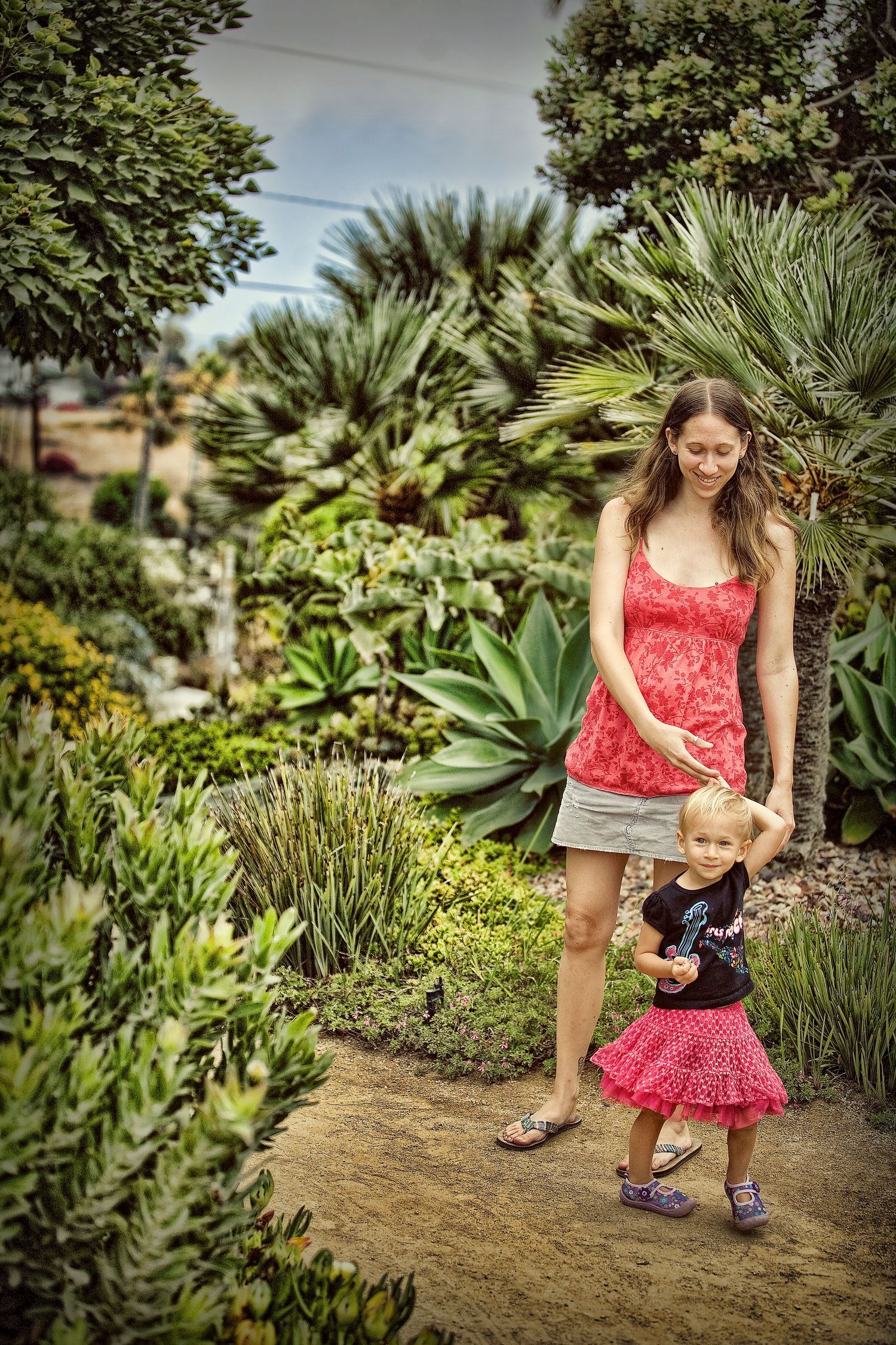 A woman in a red top guides a toddler girl in a pink skirt through a lush garden path with tropical plants.