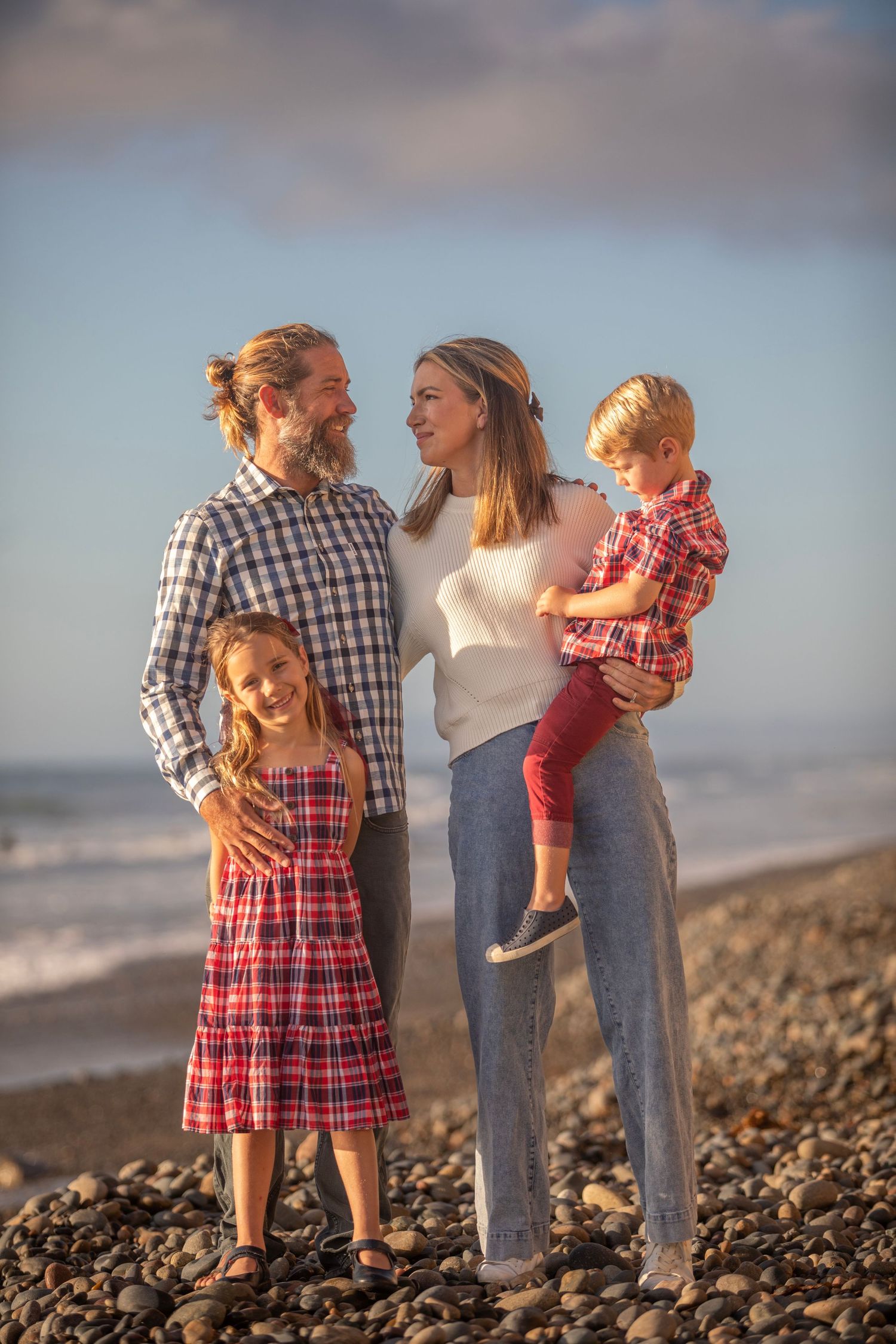 Happy family of four posing on a rocky beach, parents and two young children in matching plaid outfits at sunset.
