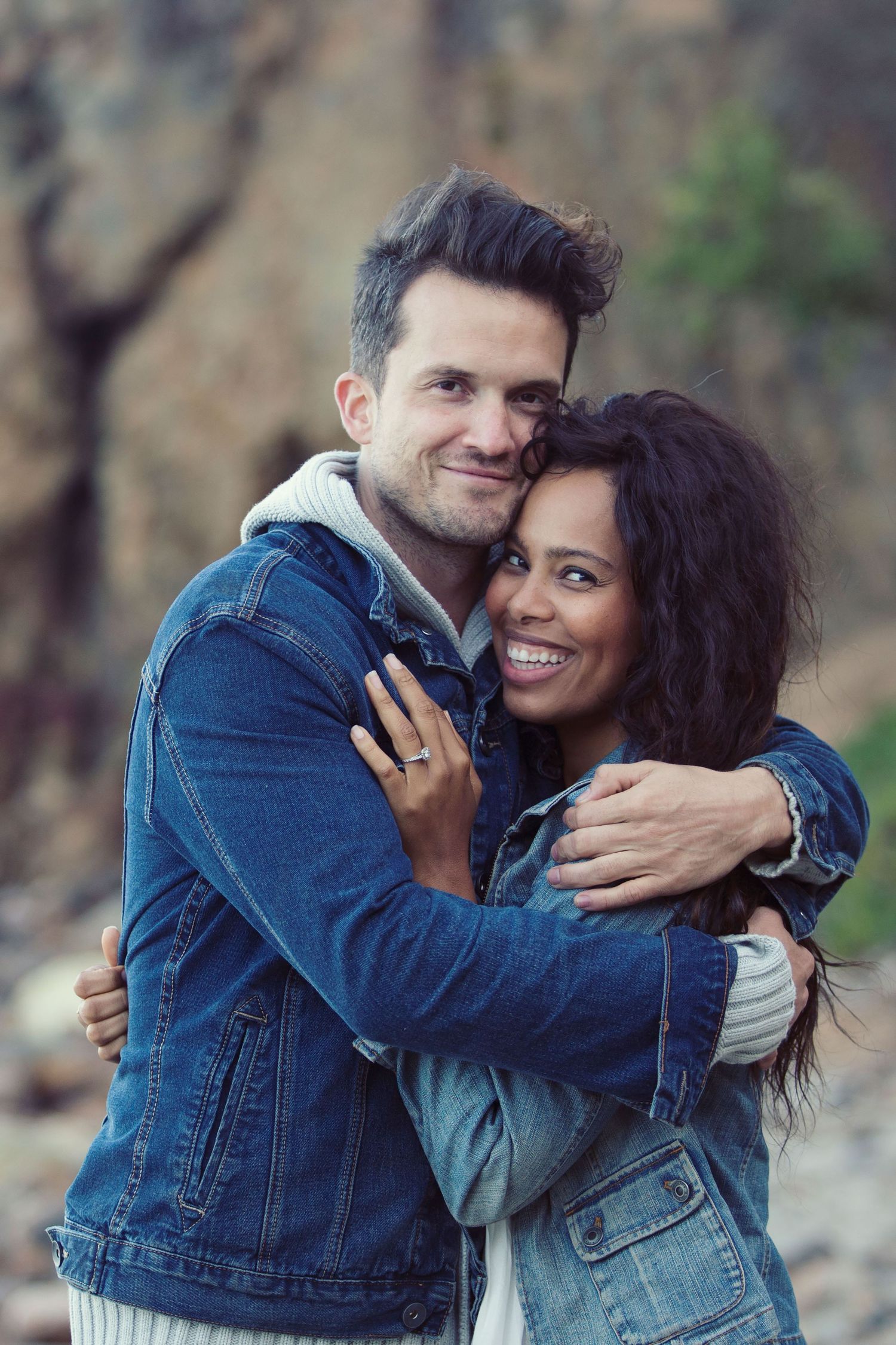 Happy interracial couple embracing and smiling outdoors near rocky cliffs in denim jackets.