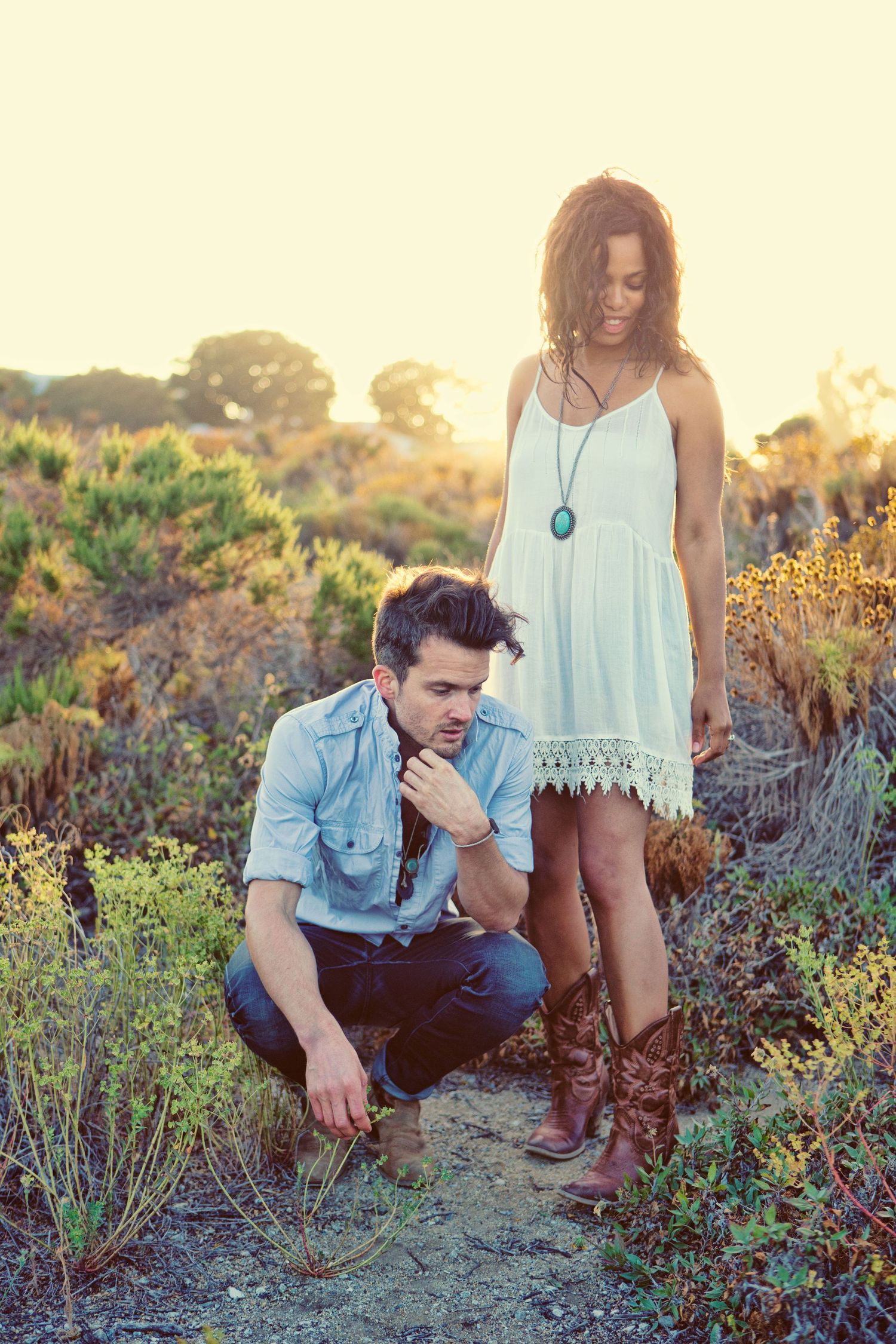 Couple in boho style outdoors at sunset, man crouching in denim shirt, woman in white lace dress with turquoise necklace.