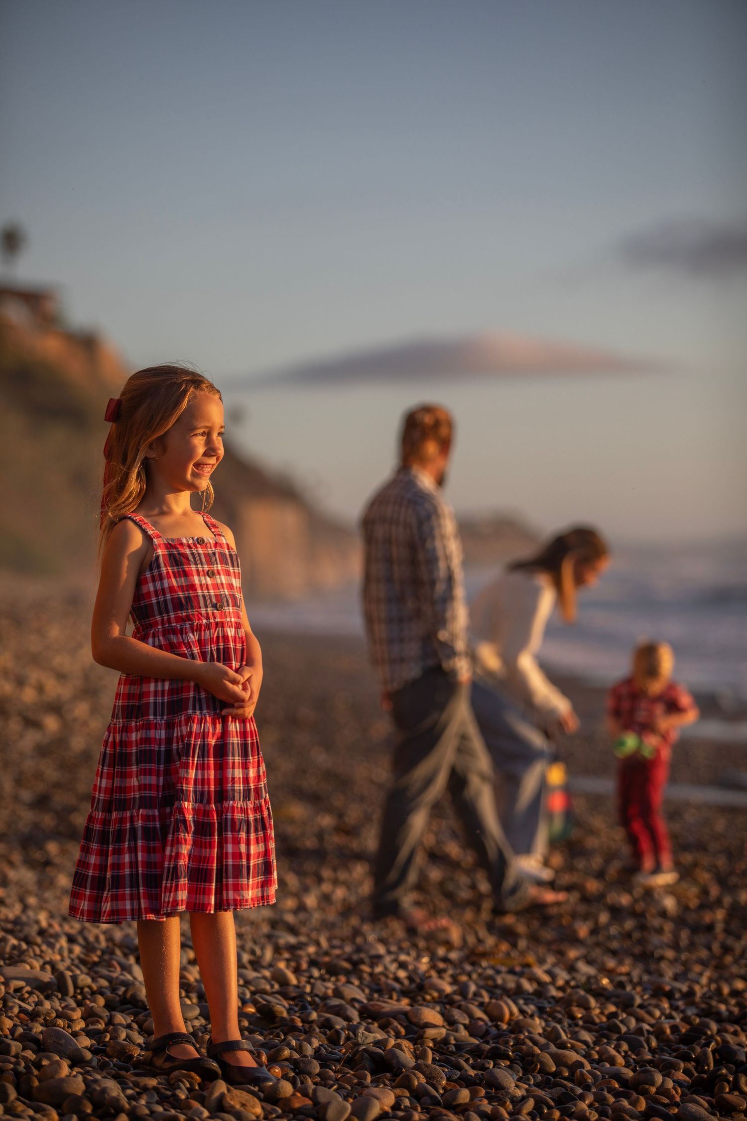 A smiling girl in a red plaid dress stands on a rocky beach at sunset with her family in the background.