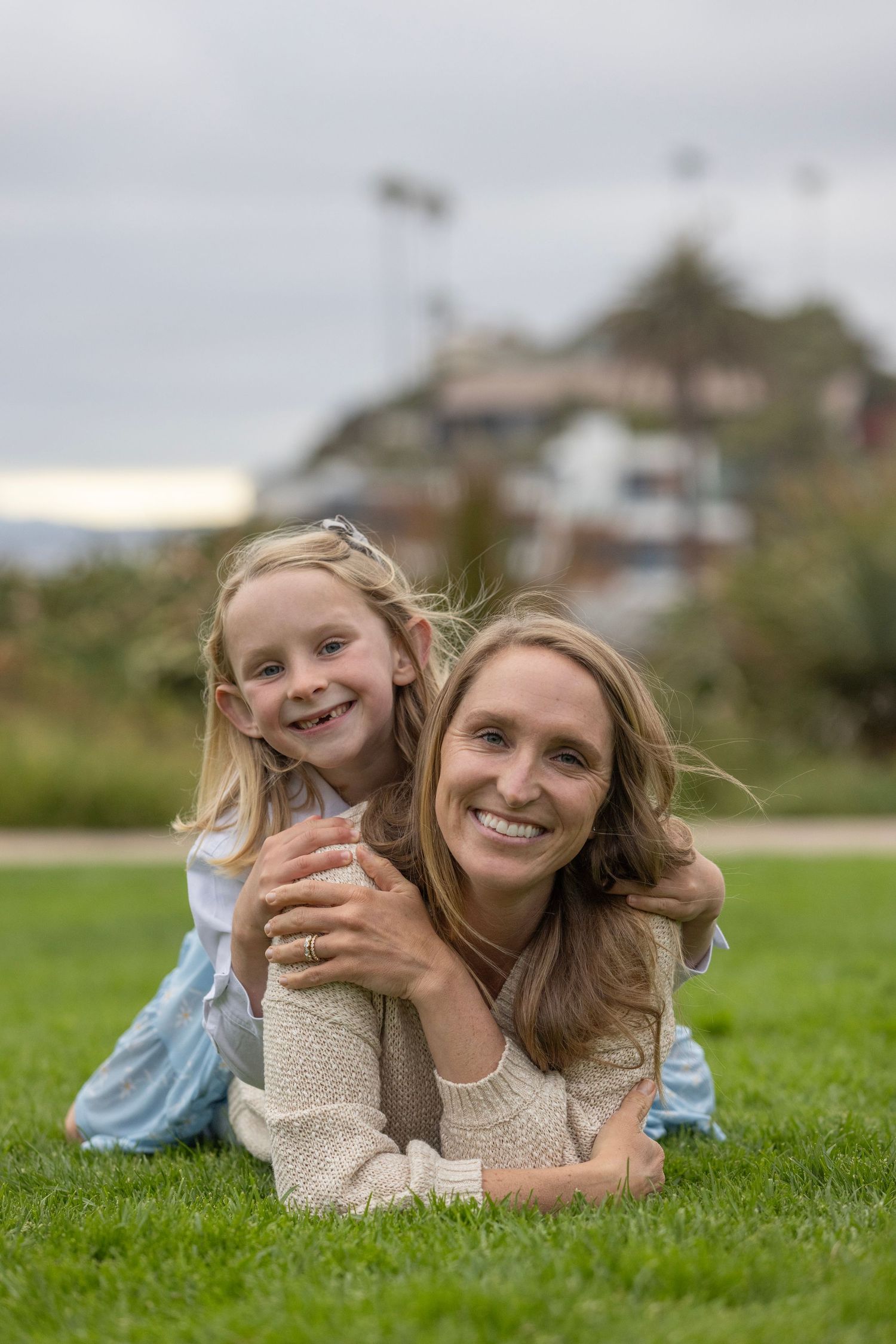 A smiling mother and daughter lying on green grass, with the young girl hugging her mom from behind outdoors.