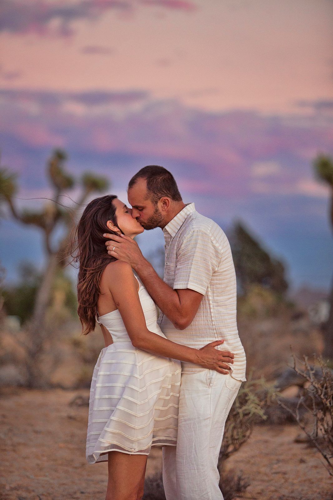Couple sharing a romantic kiss in a desert landscape at sunset, both dressed in white, with Joshua trees and colorful sky.