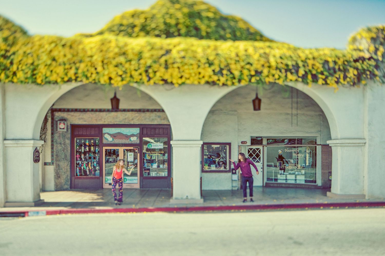 People walking in front of a white arched building with lush green ivy on the roof in a sunny town.
