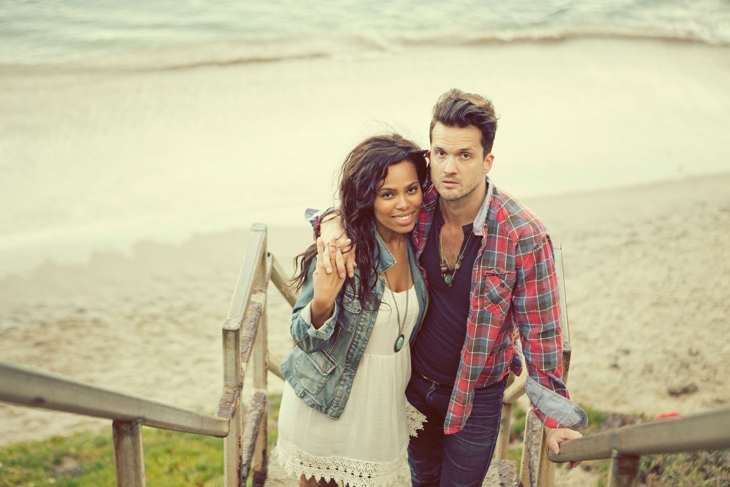 A couple poses on beach stairs, woman in denim jacket holding keys, man in plaid shirt, sandy shore behind them.