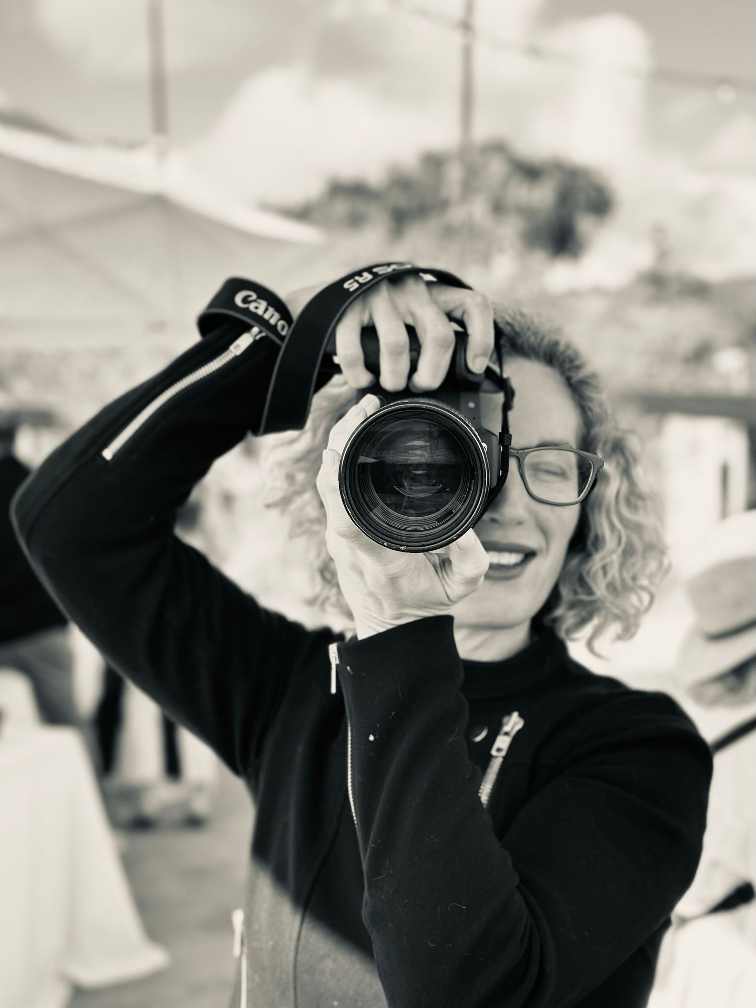 Black and white photo of a smiling woman with curly hair holding a Canon camera up to take a picture outdoors.