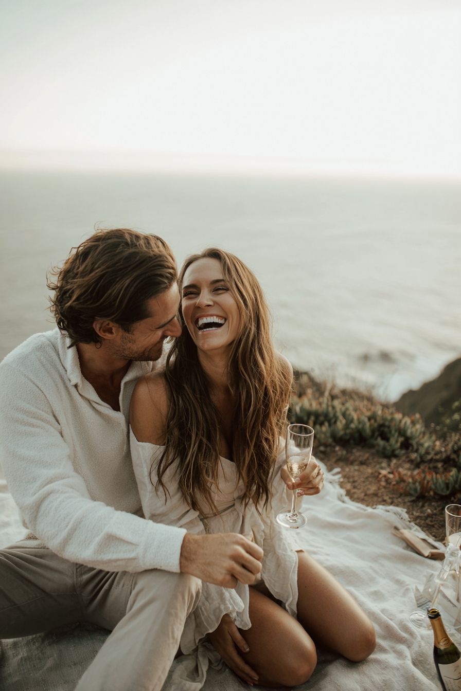 Couple laughing and drinking champagne on a coastal cliff at sunset, enjoying a romantic outdoor picnic by the ocean.