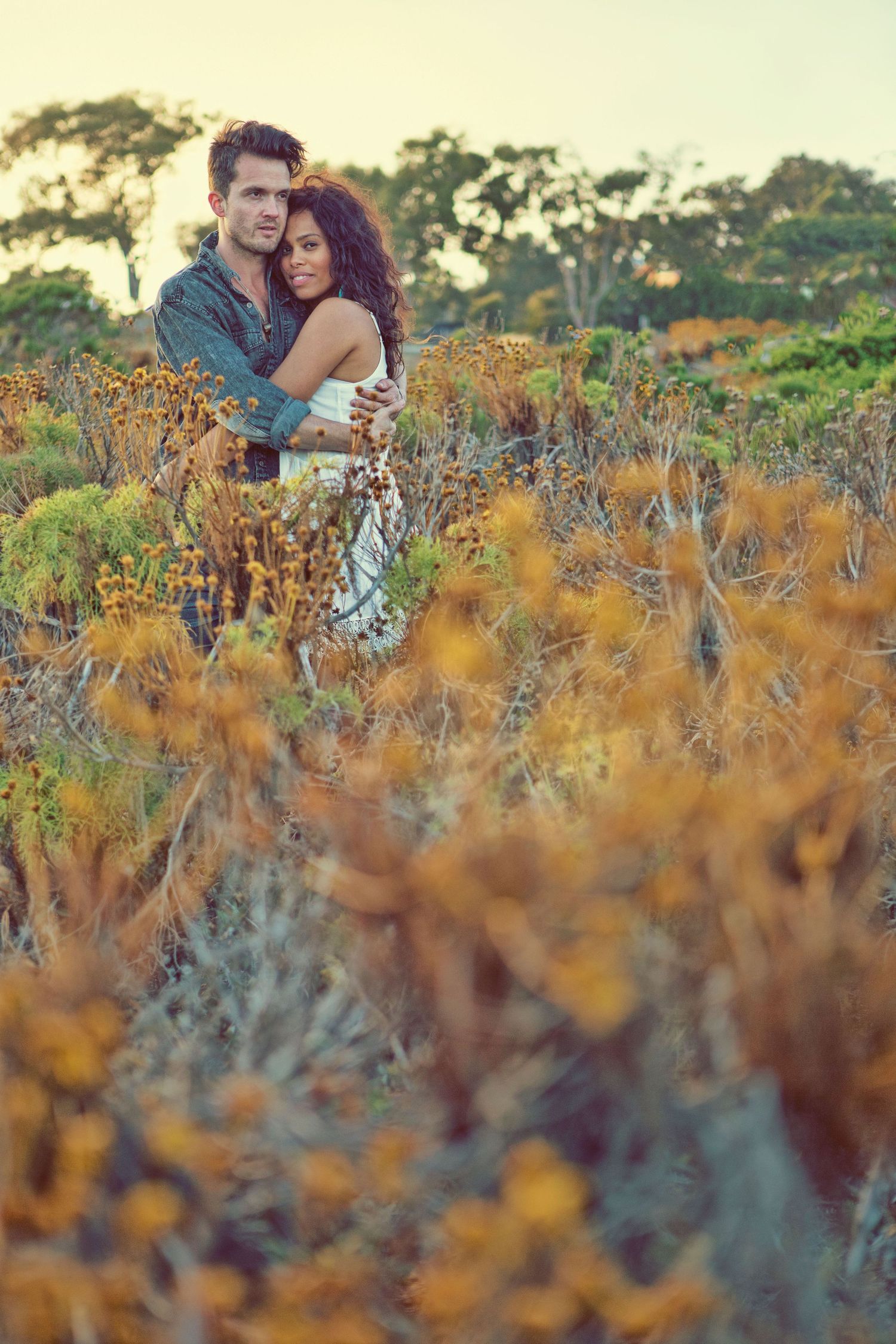 Romantic couple embracing in a field of yellow wildflowers at sunset, surrounded by lush greenery.