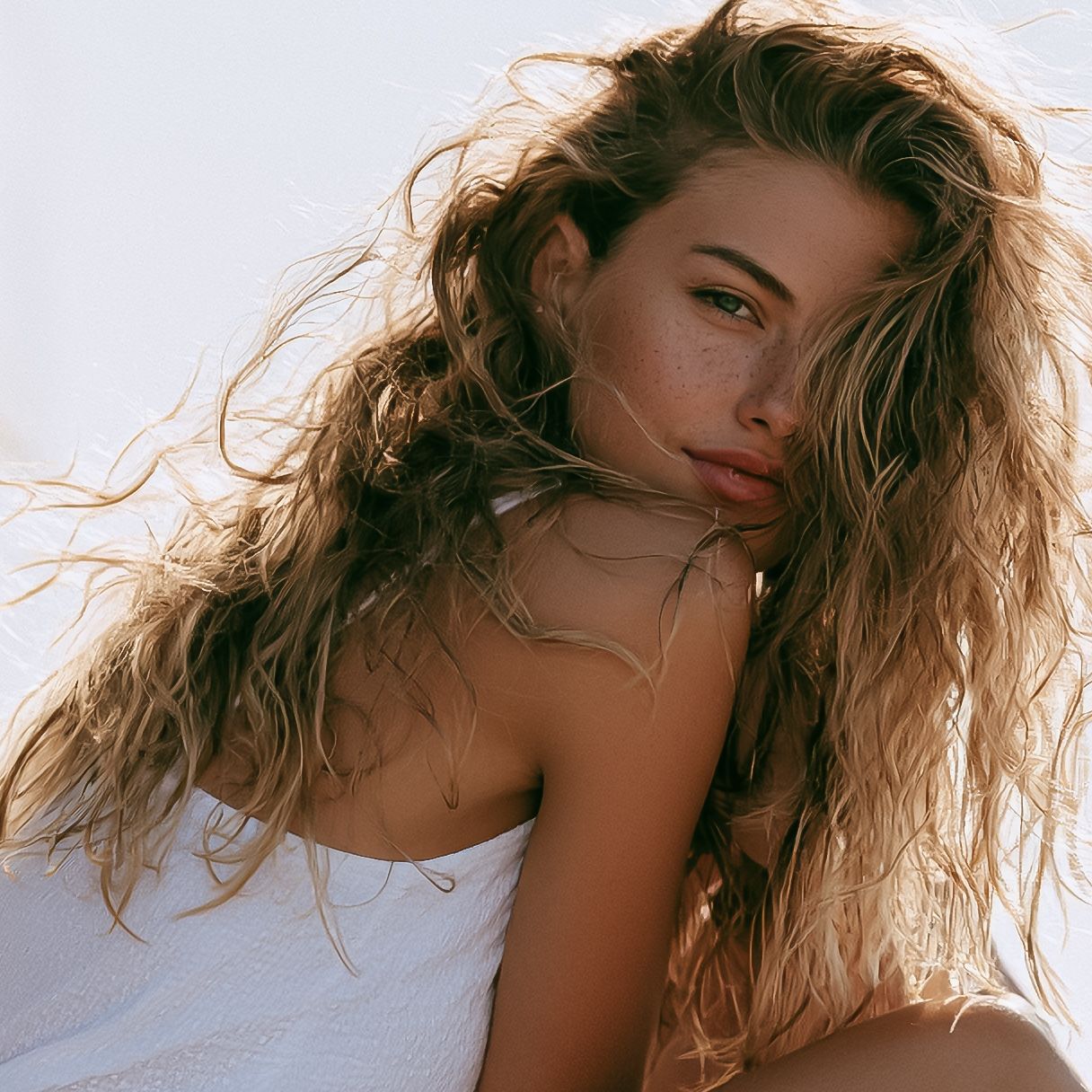Young woman with long wavy sun-kissed hair and freckles posing outdoors in bright natural light wearing a white top.
