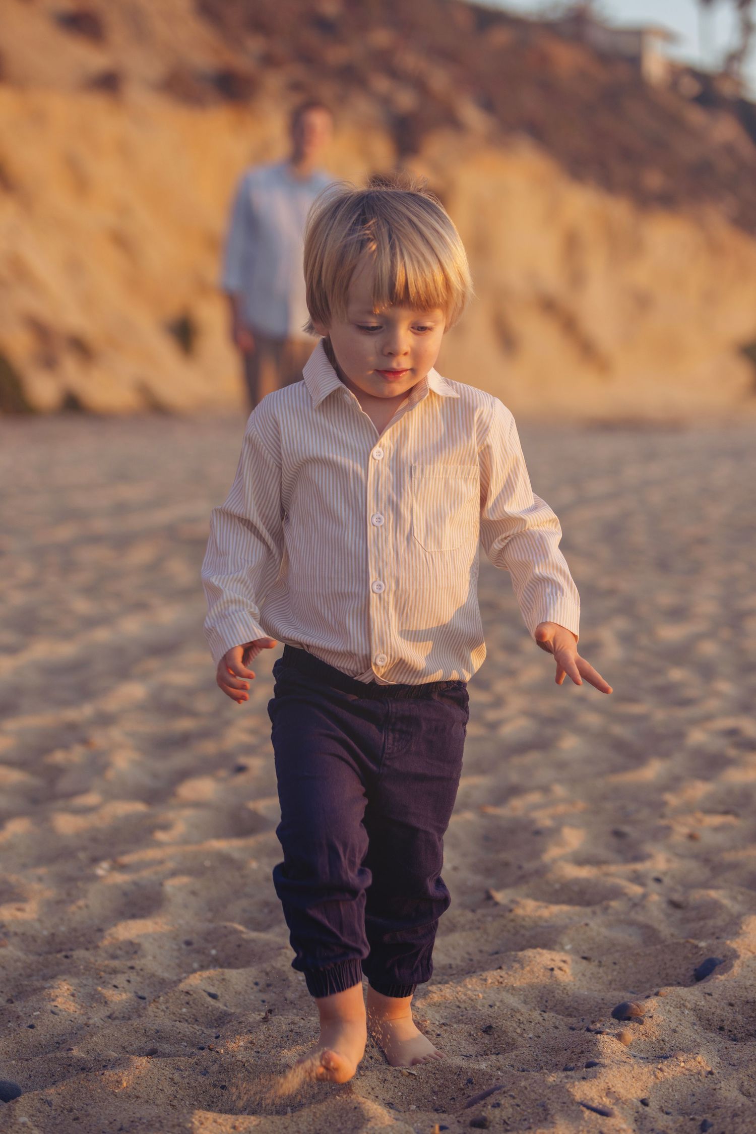 A young blond boy in a white shirt and navy pants walks barefoot on a sandy beach at golden hour.