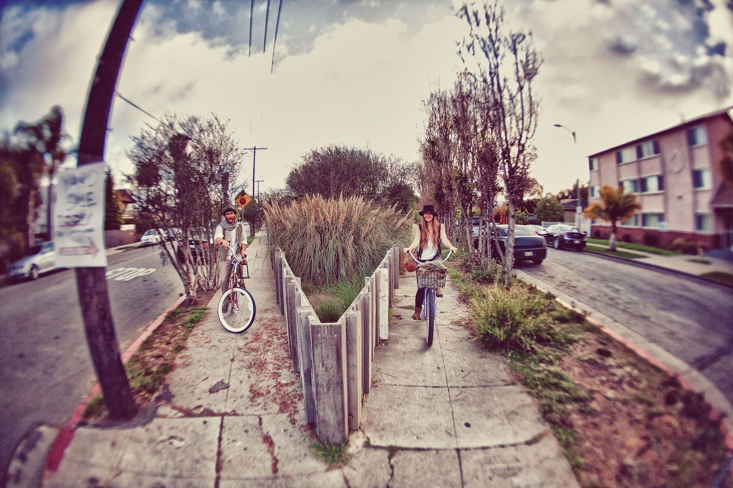 Fisheye lens photo of a urban sidewalk with cyclists riding past a landscaped median strip on a cloudy day.