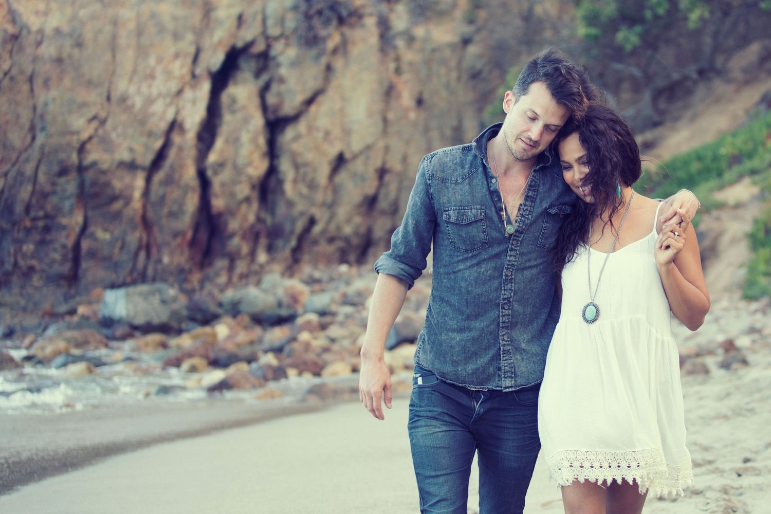 Romantic couple walking on a sandy beach near rocky cliffs, man in denim shirt with arm around woman in white dress.