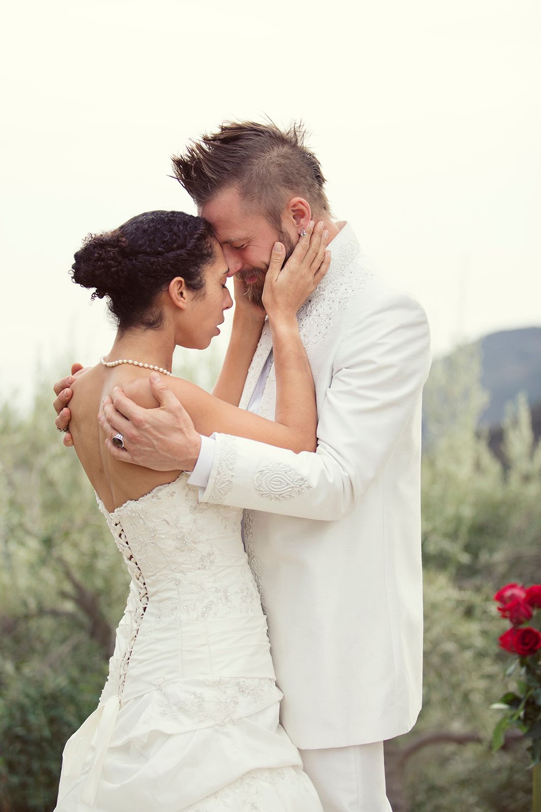 Romantic bride and groom in white embrace tenderly outdoors with red roses and lush greenery in background.