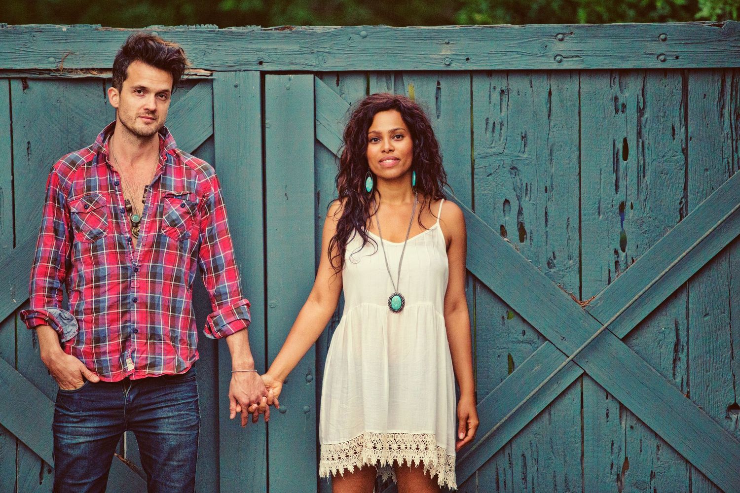 A couple holds hands in front of a rustic teal barn door, man in plaid shirt and woman in white dress.