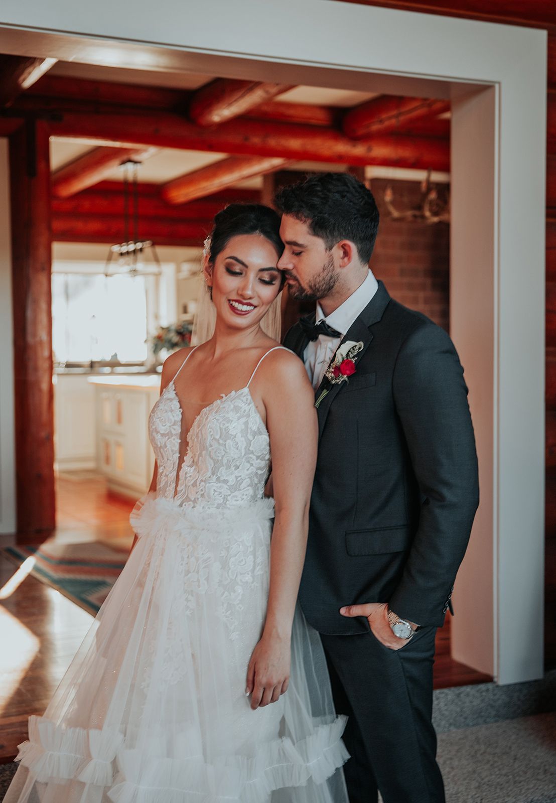 Bride in lace gown and groom in dark suit share romantic moment, groom kissing bride's cheek indoors.