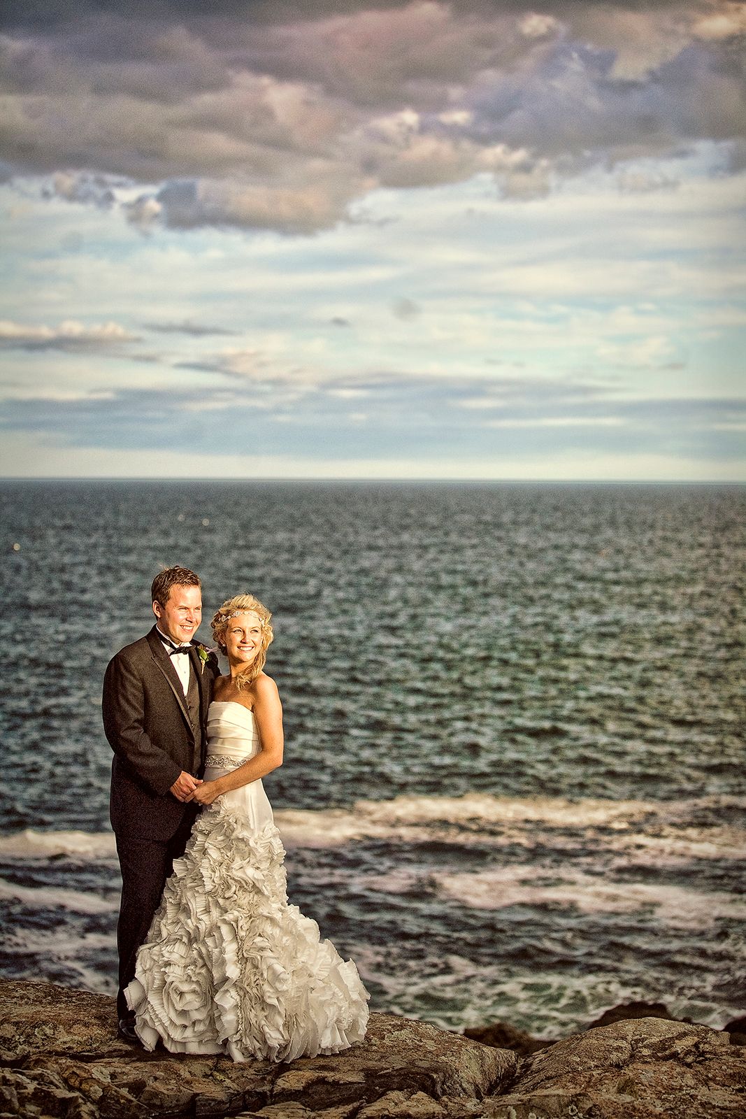 Bride and groom posing on rocky shoreline with dramatic ocean and cloudy sky backdrop at sunset wedding photo.