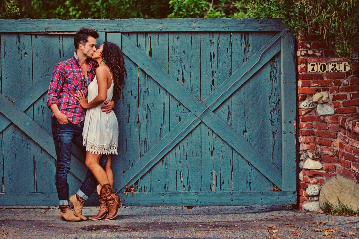Romantic couple kissing against a rustic turquoise wooden gate beside a brick wall outdoors.