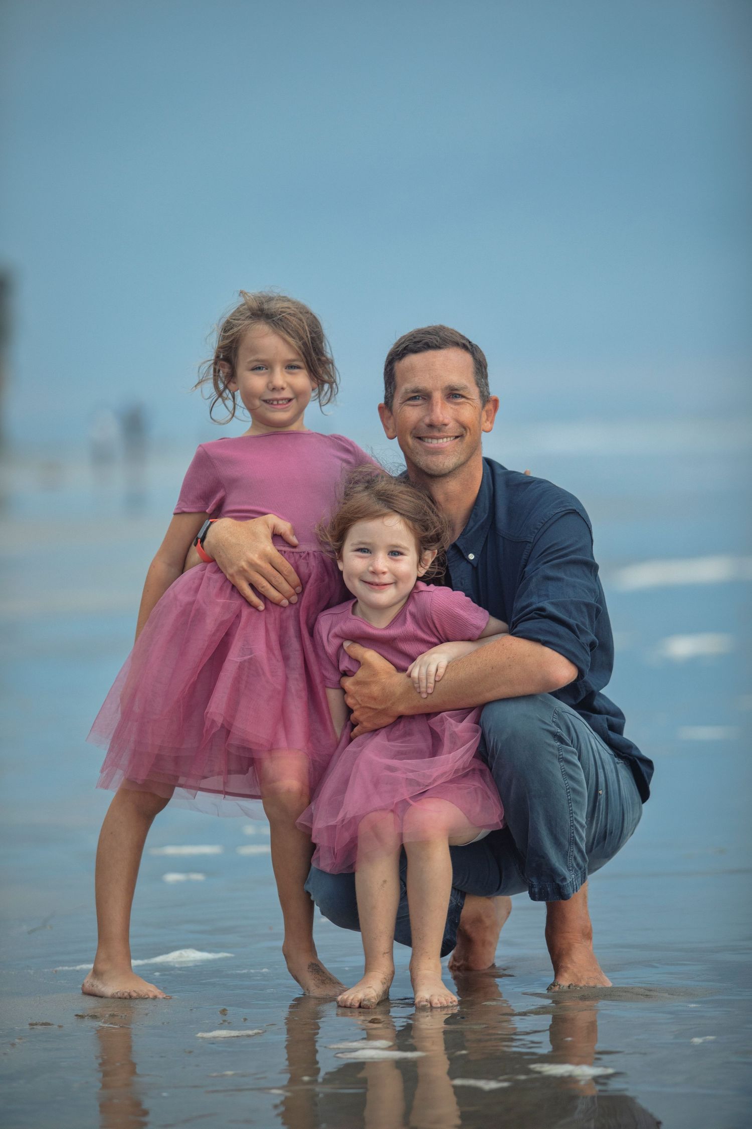 A smiling father crouches on a wet beach with his two young daughters in matching pink tulle dresses.