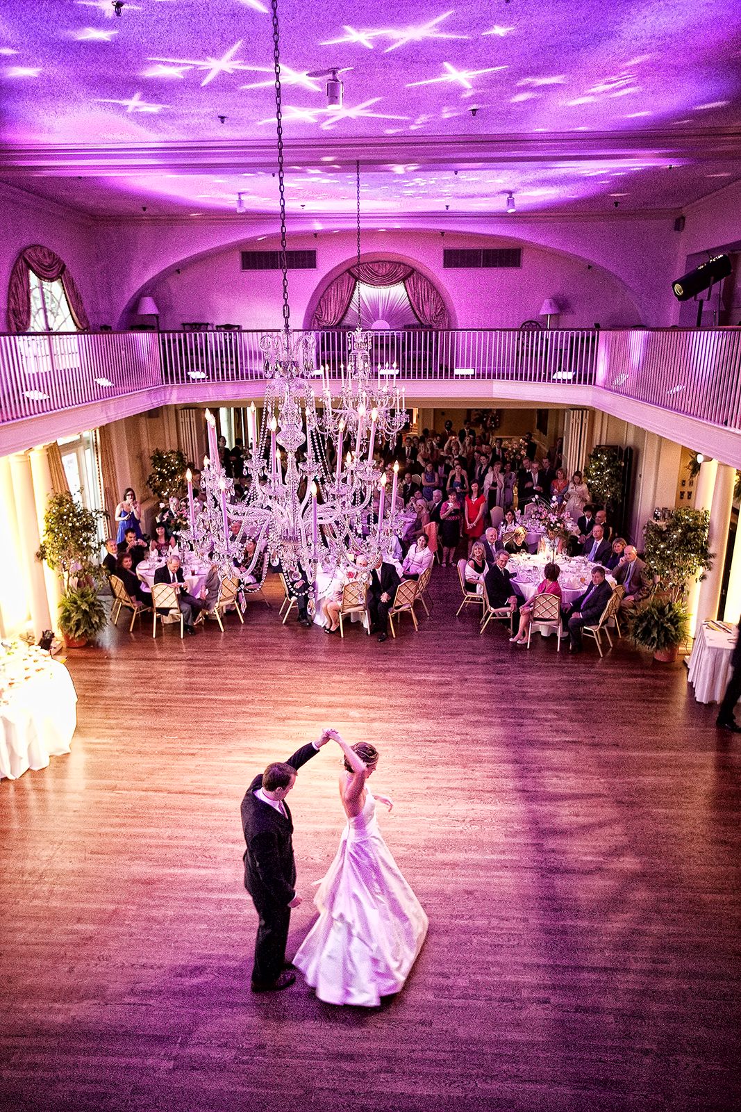 Bride and groom share first dance under crystal chandelier in elegant ballroom with purple uplighting and guests seated around dance floor.