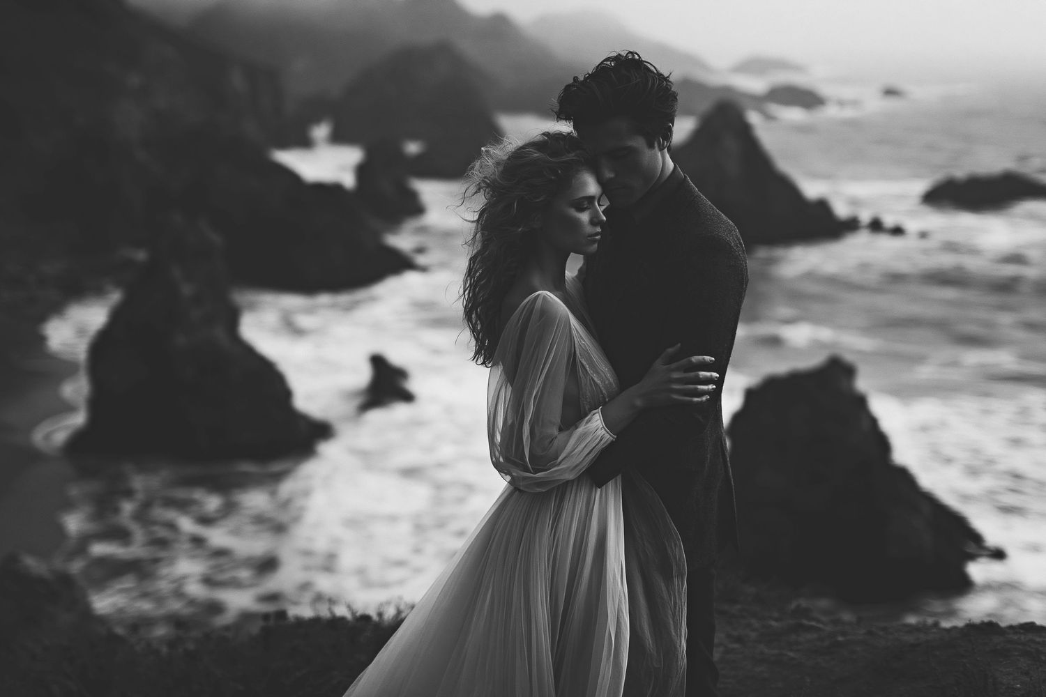 Black and white photo of a romantic couple embracing on a rocky coastal cliff with dramatic ocean waves in the background.