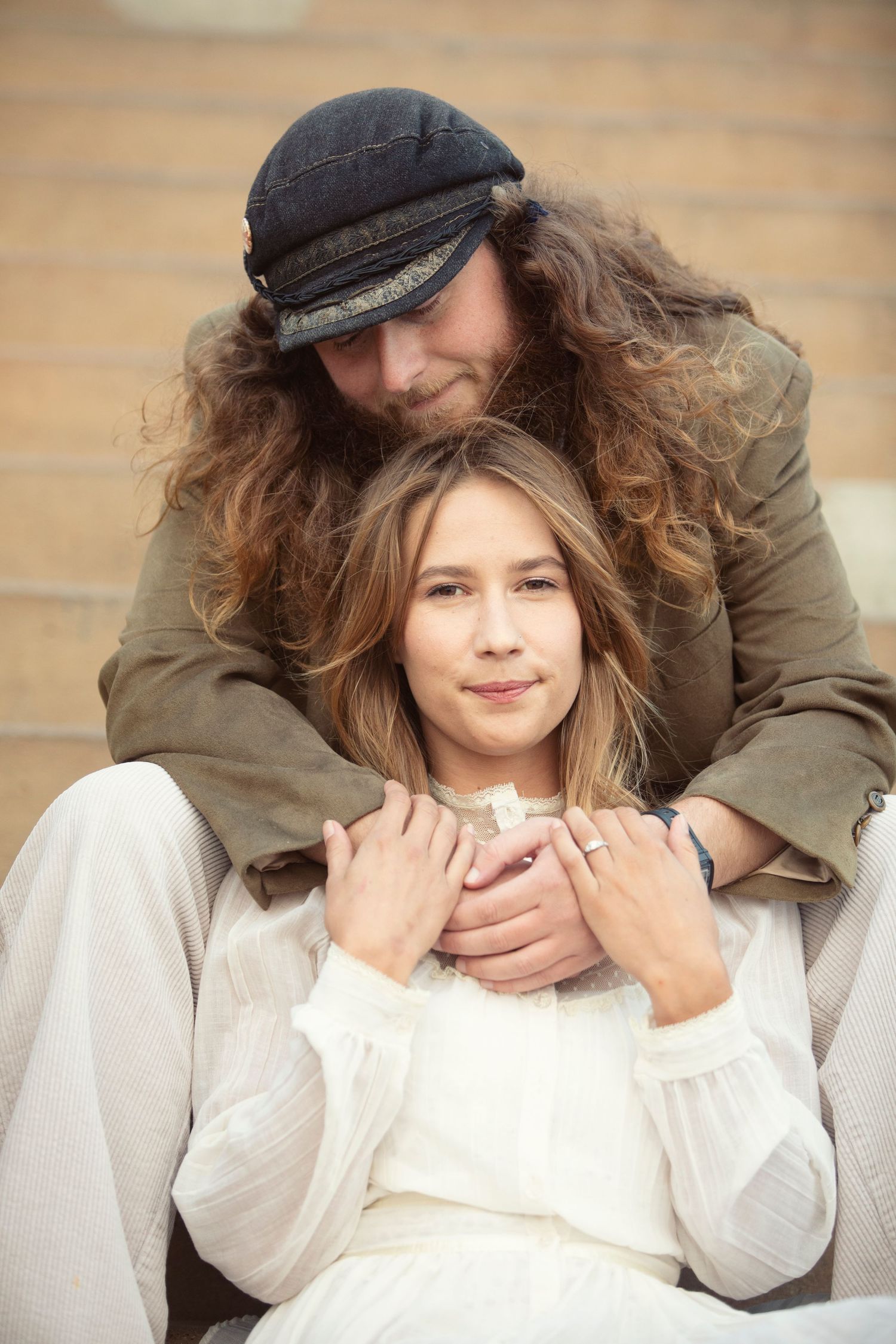 Couple with long curly hair sitting together outdoors, woman in white dress, man in dark cap embracing her from behind.