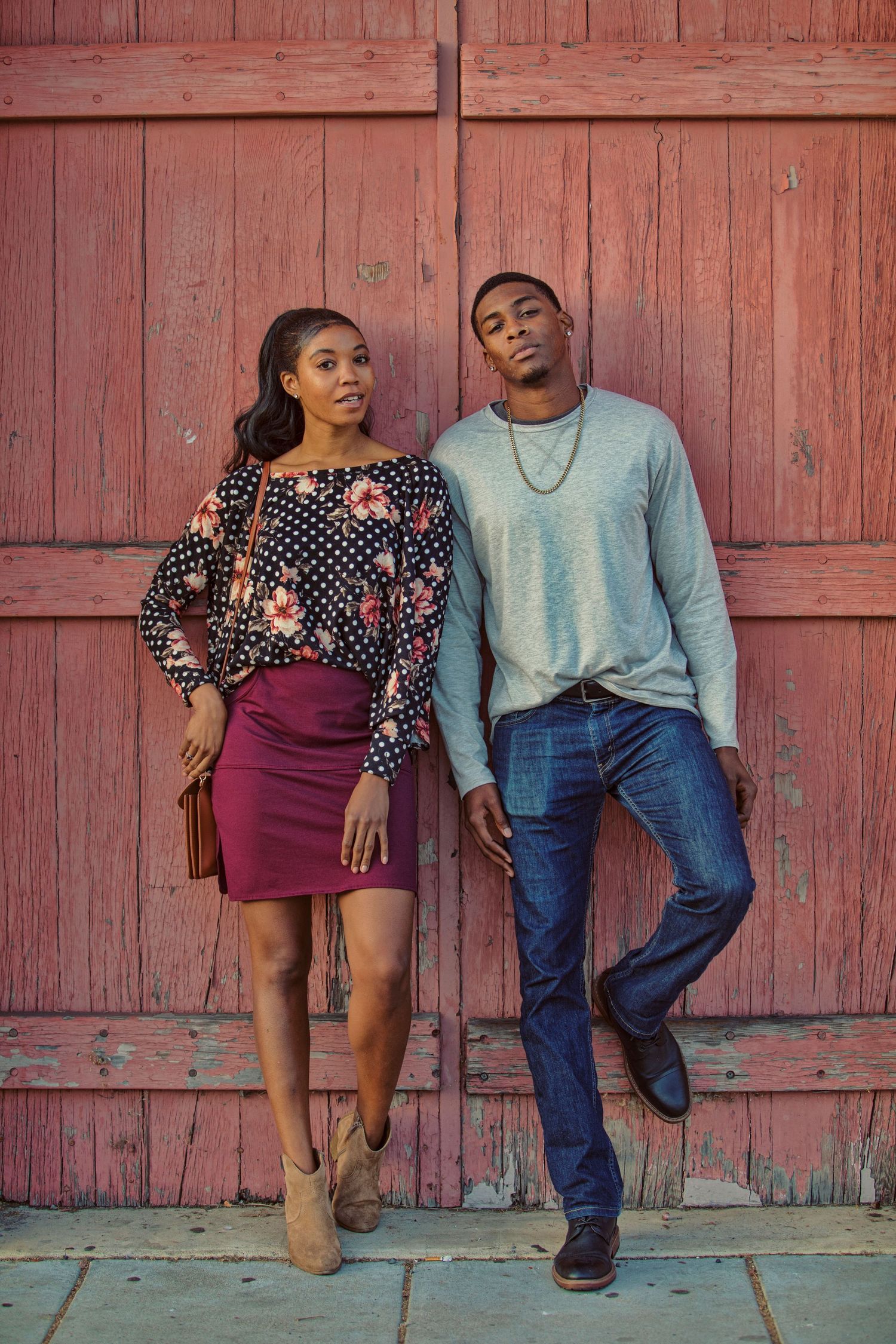 Young Black couple posing confidently against a rustic red barn door, dressed in stylish casual outfits.