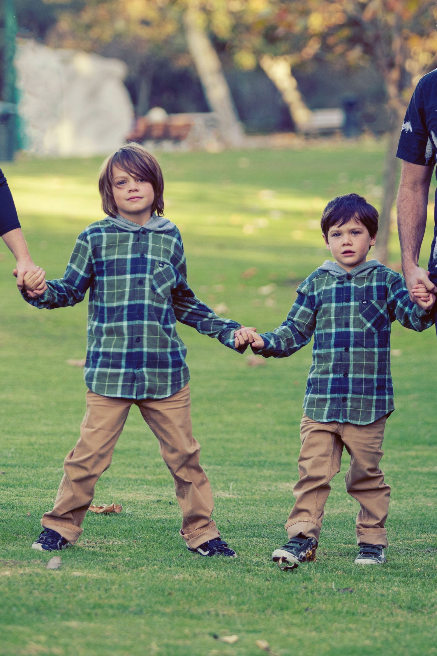 Two young boys in matching plaid shirts and khaki pants holding hands outdoors on green grass.
