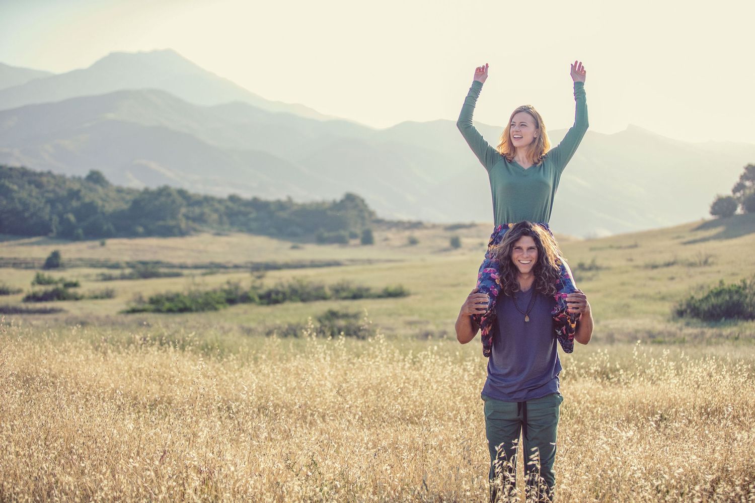 Woman sitting on man's shoulders with arms raised in a golden field with mountain backdrop on a sunny day.