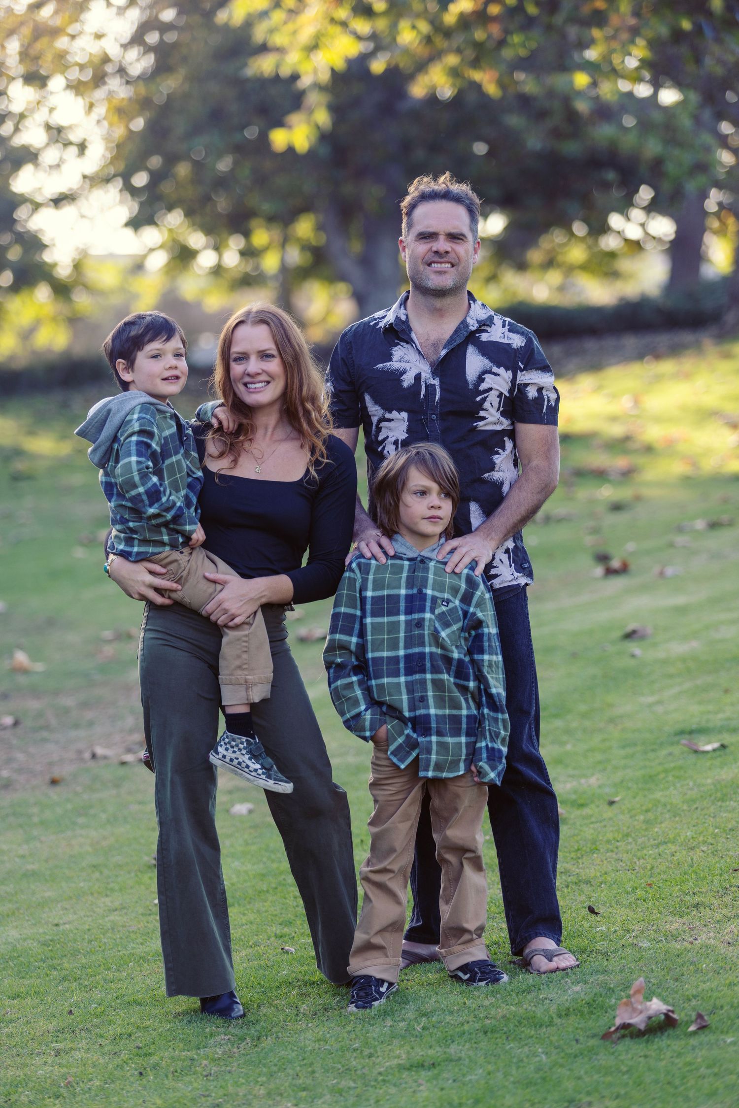 Family of four posing outdoors on green grass surrounded by autumn trees in a warm, natural setting.