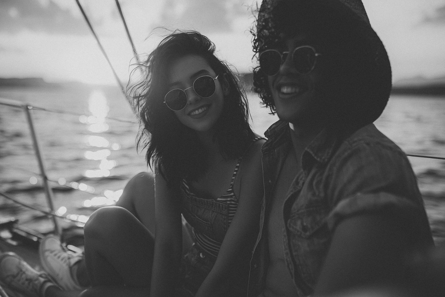 Black and white photo of two smiling women wearing sunglasses on a sailboat at sunset.
